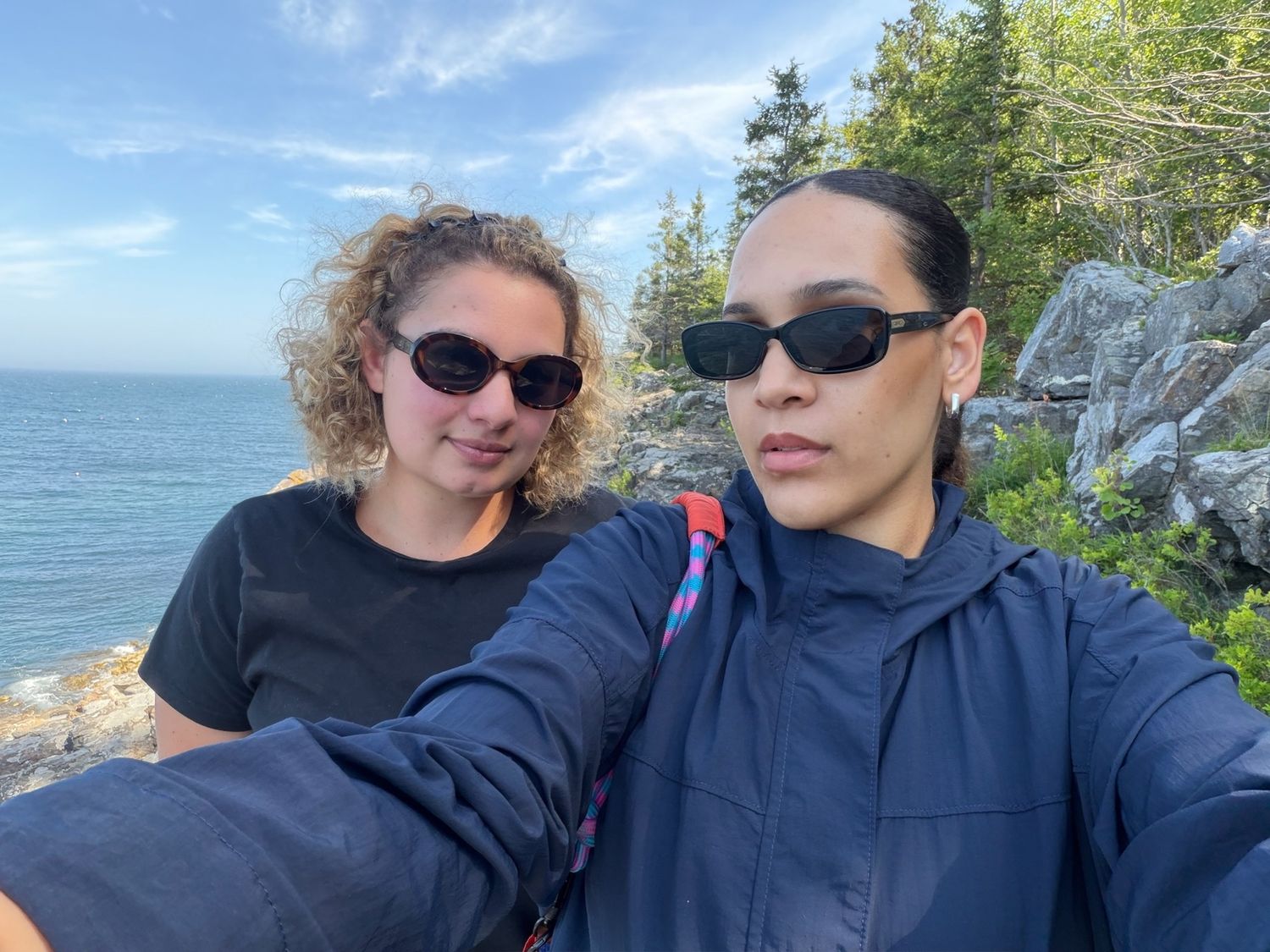 Two friends wearing sunglasses take a selfie on a rocky coastal trail with blue ocean and trees in the background.