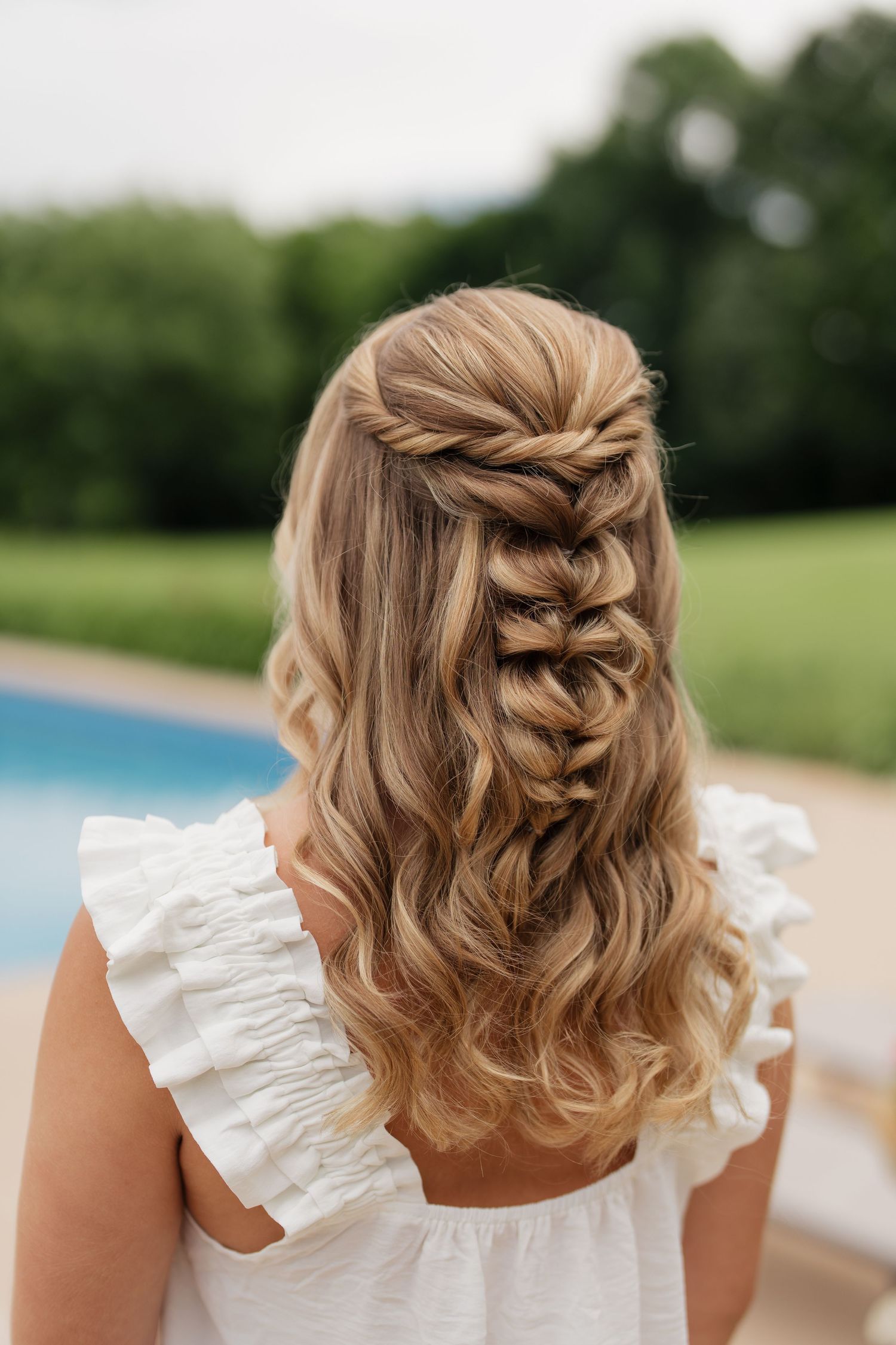 Back view of elegant half-up braided hairstyle with loose waves on blonde hair, outdoors near a pool.