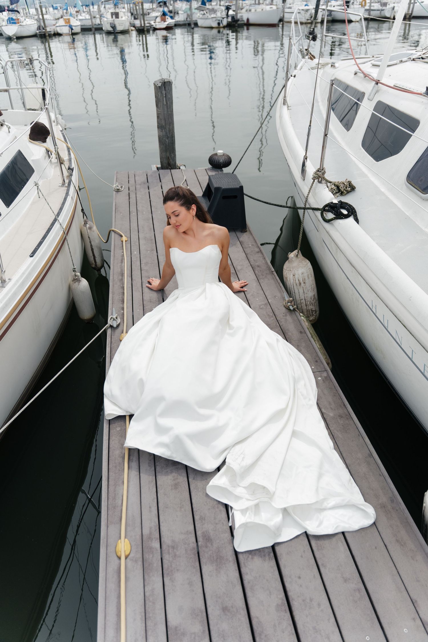 Bride in a white ball gown sitting on a marina dock surrounded by sailboats, with her dress spread elegantly around her.