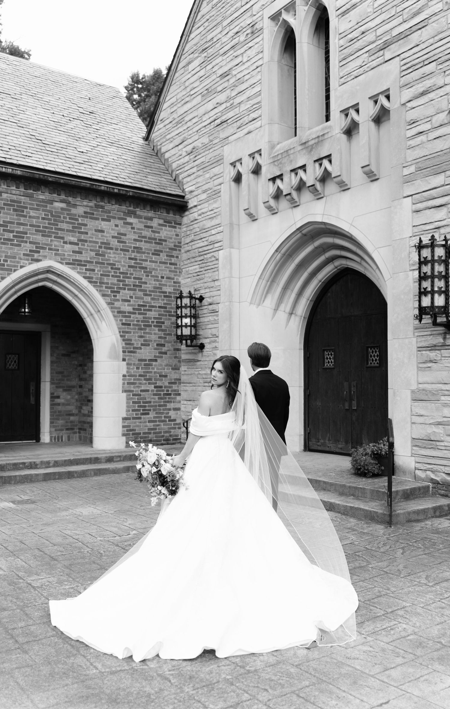Black and white photo of bride and groom standing outside a stone church, bride wearing off-shoulder gown with long veil.
