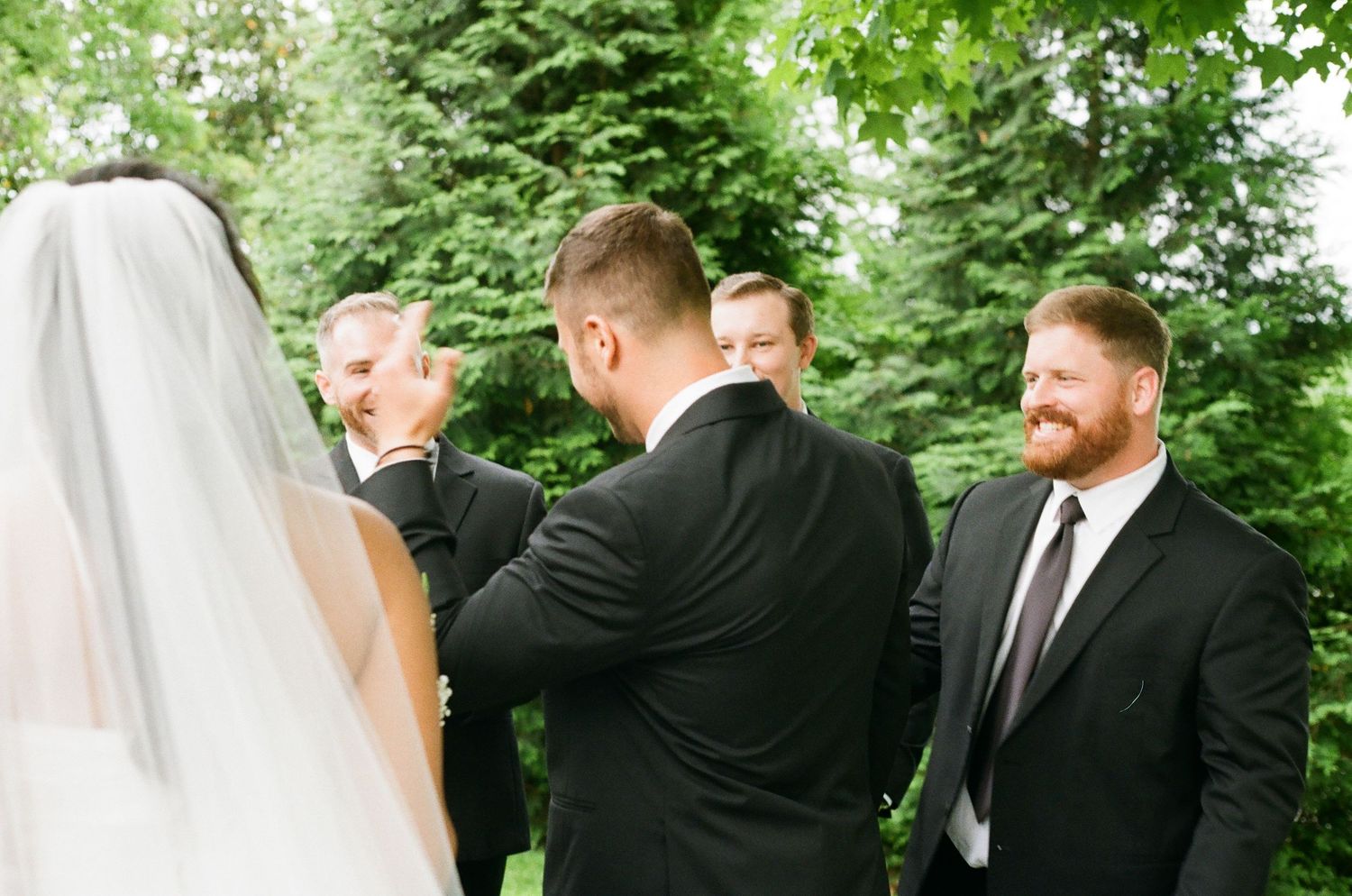 Groomsmen in black suits gather outdoors during a wedding ceremony, with a bride in a white veil visible in the foreground.