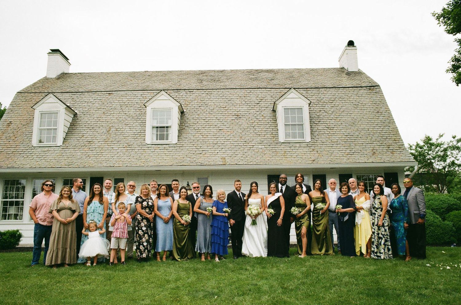 Large wedding party group photo in front of a charming white cottage with a shingled roof.
