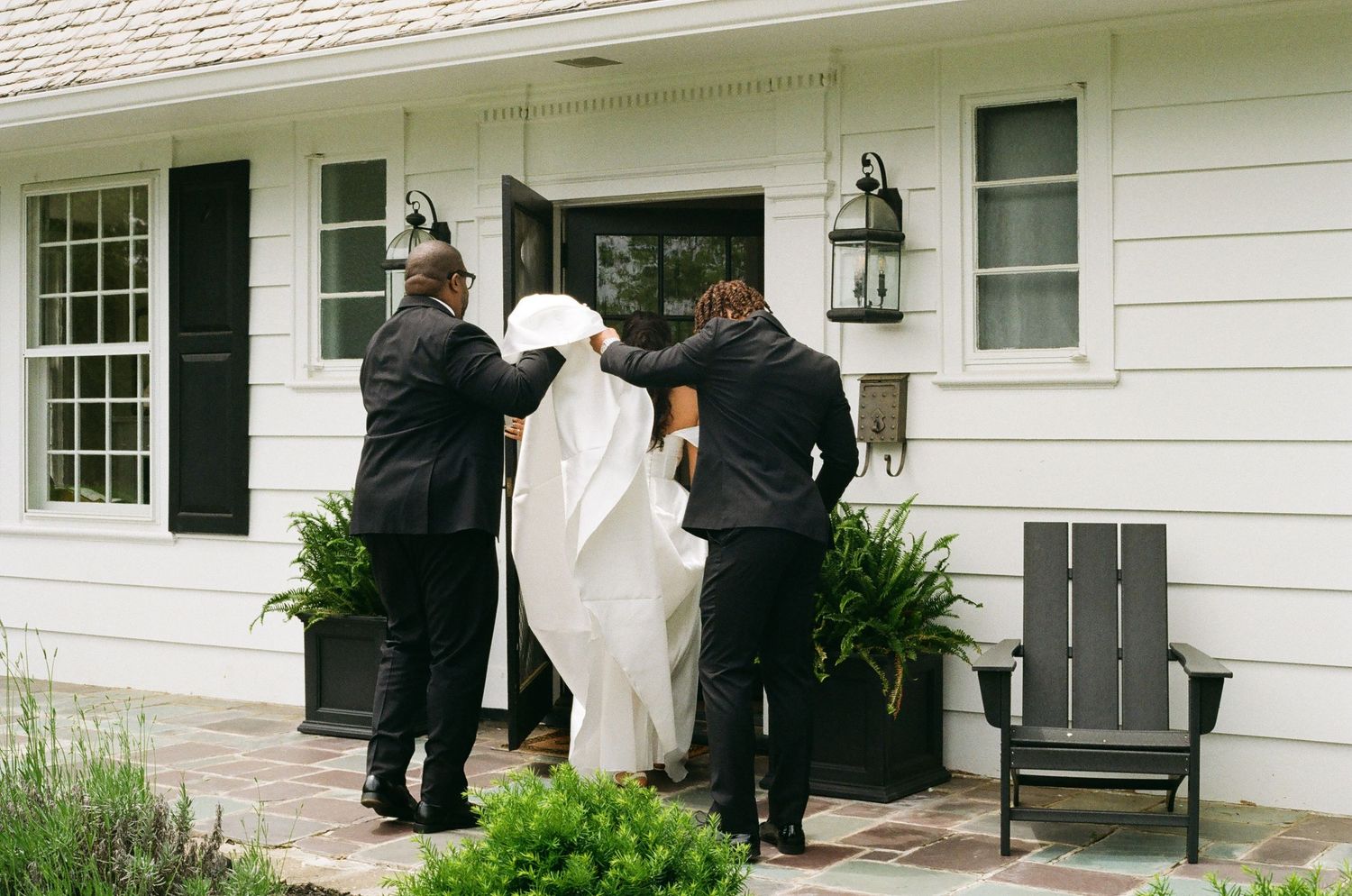 Two groomsmen help a bride in a white gown enter a white house with black shutters on her wedding day.