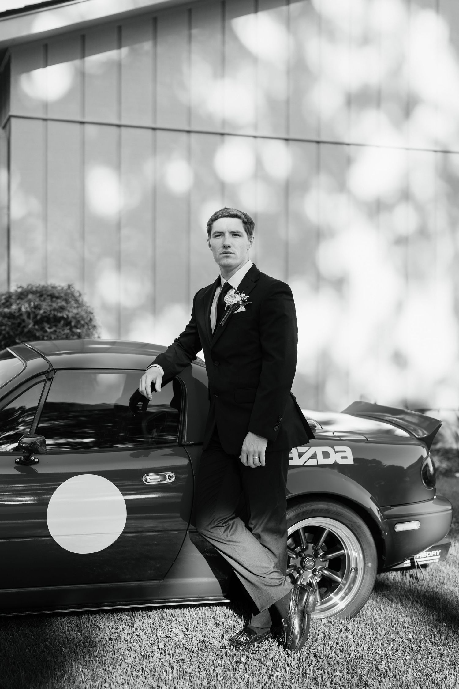 Groom in black suit with boutonniere leaning against a Mazda sports car in elegant black and white wedding photo.