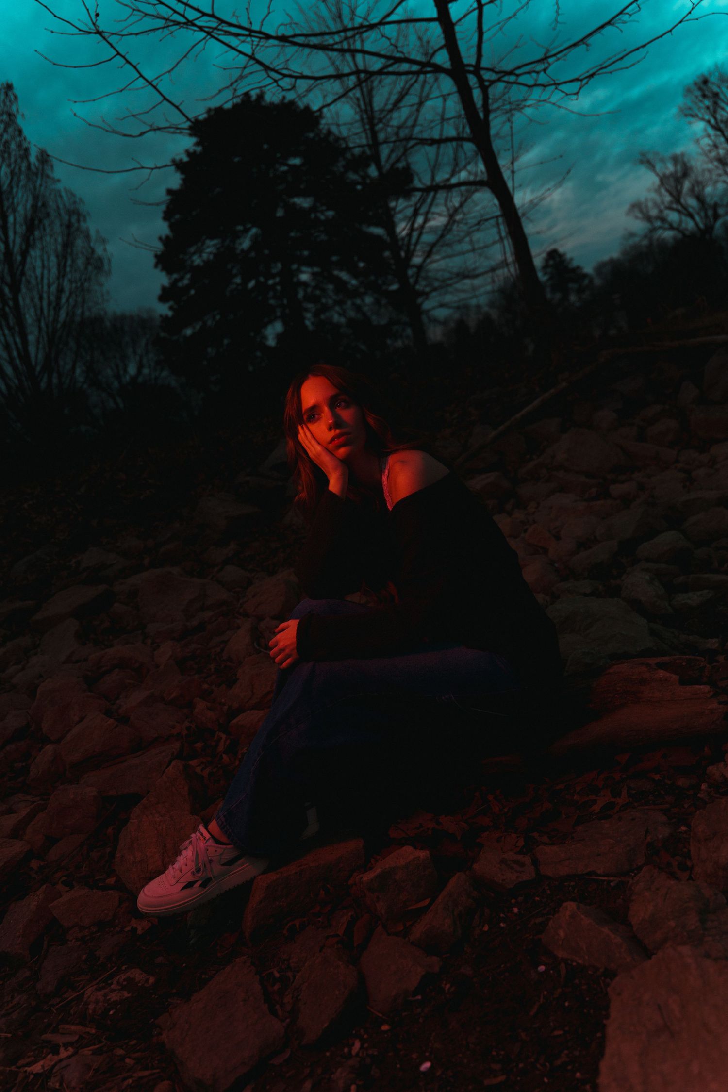 Moody nighttime portrait on rocky terrain with dark silhouetted trees and a teal-colored sky in the background.