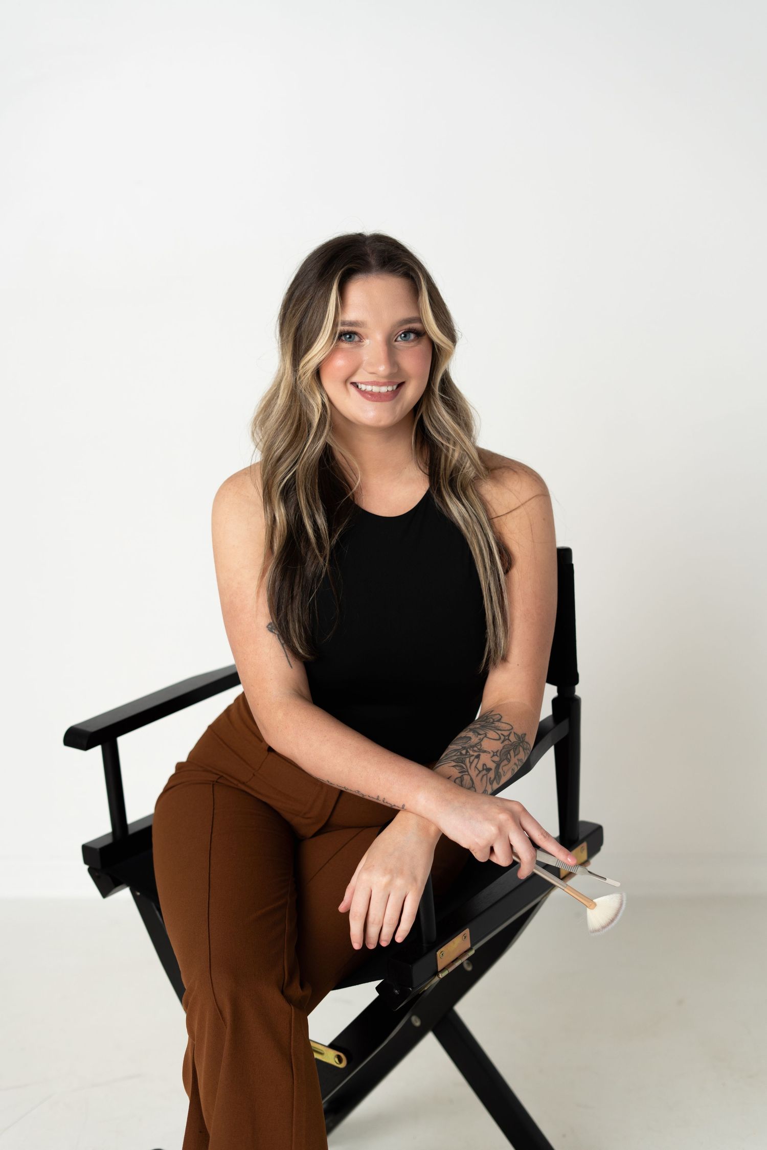 Professional lifestyle portrait of someone sitting on a black director's chair wearing a black sleeveless top and brown pants.