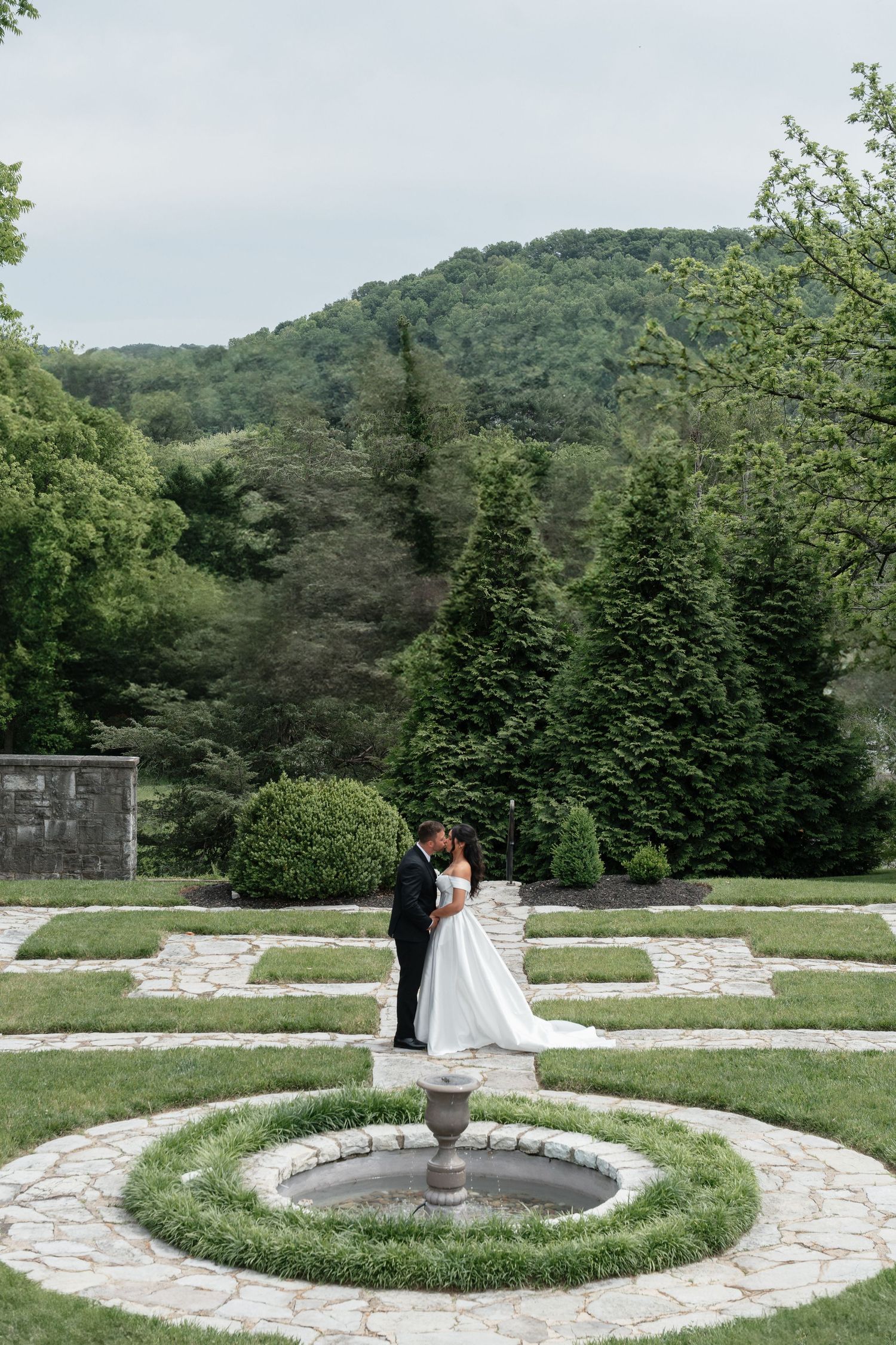 Bride and groom kissing near a stone fountain in an elegant garden with lush green trees and rolling hills backdrop.
