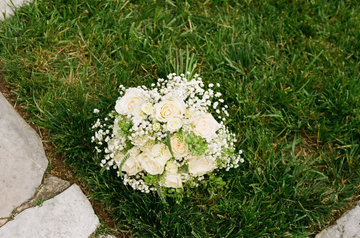 White rose and baby's breath bridal bouquet resting on lush green grass beside a stone path.