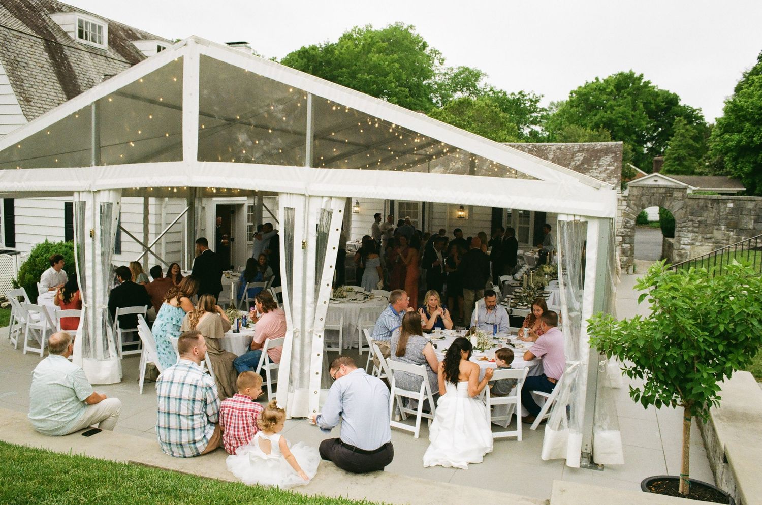 Outdoor wedding reception under a white tent with guests dining on a patio surrounded by lush greenery.