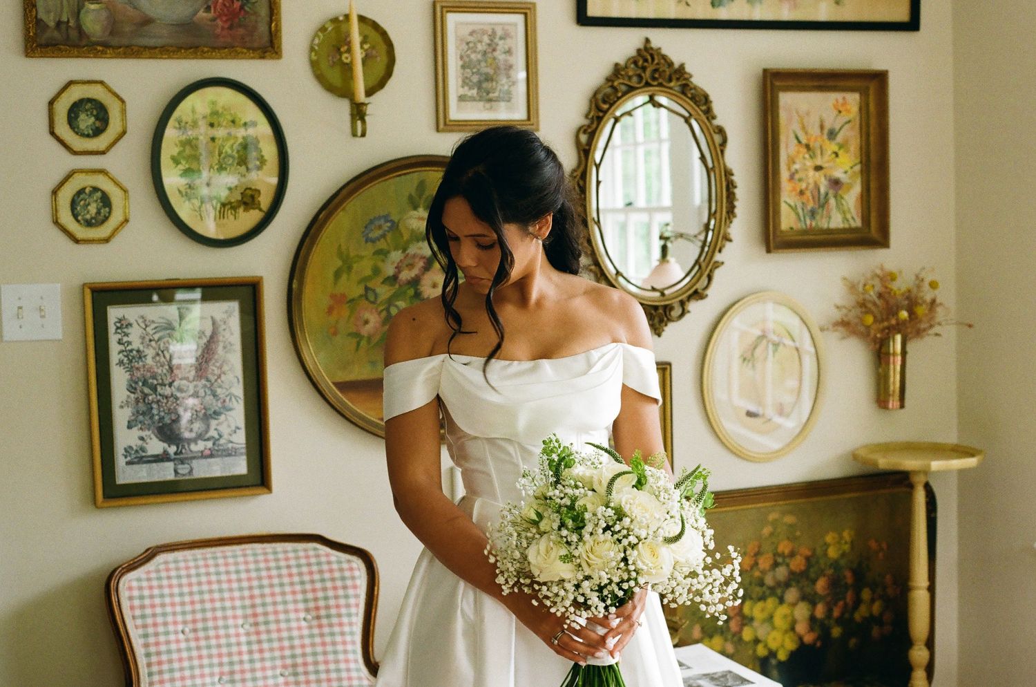 Bride in off-shoulder white gown holds white bouquet, standing before vintage mirror gallery wall in elegant room.