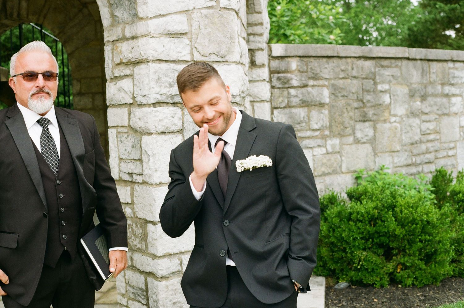 Groom in black suit with white boutonniere smiling outside stone church on wedding day.