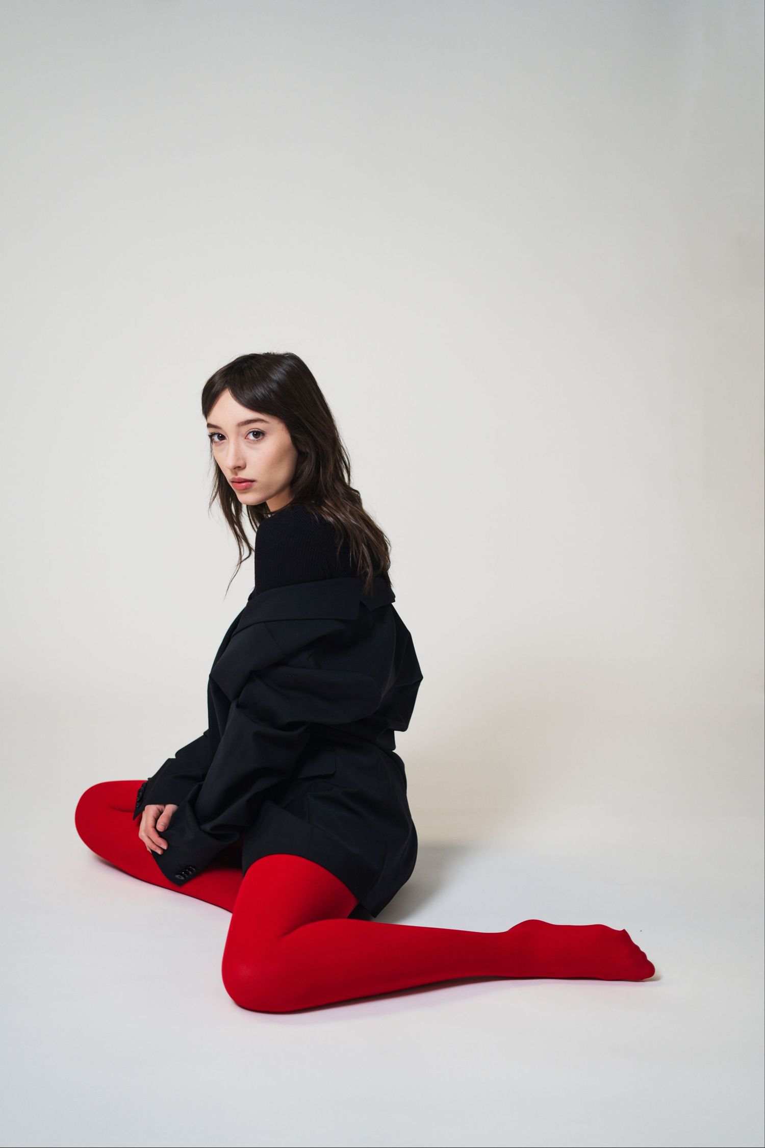 Person wearing dark clothing and red tights poses expressively in minimalist setting with white background.