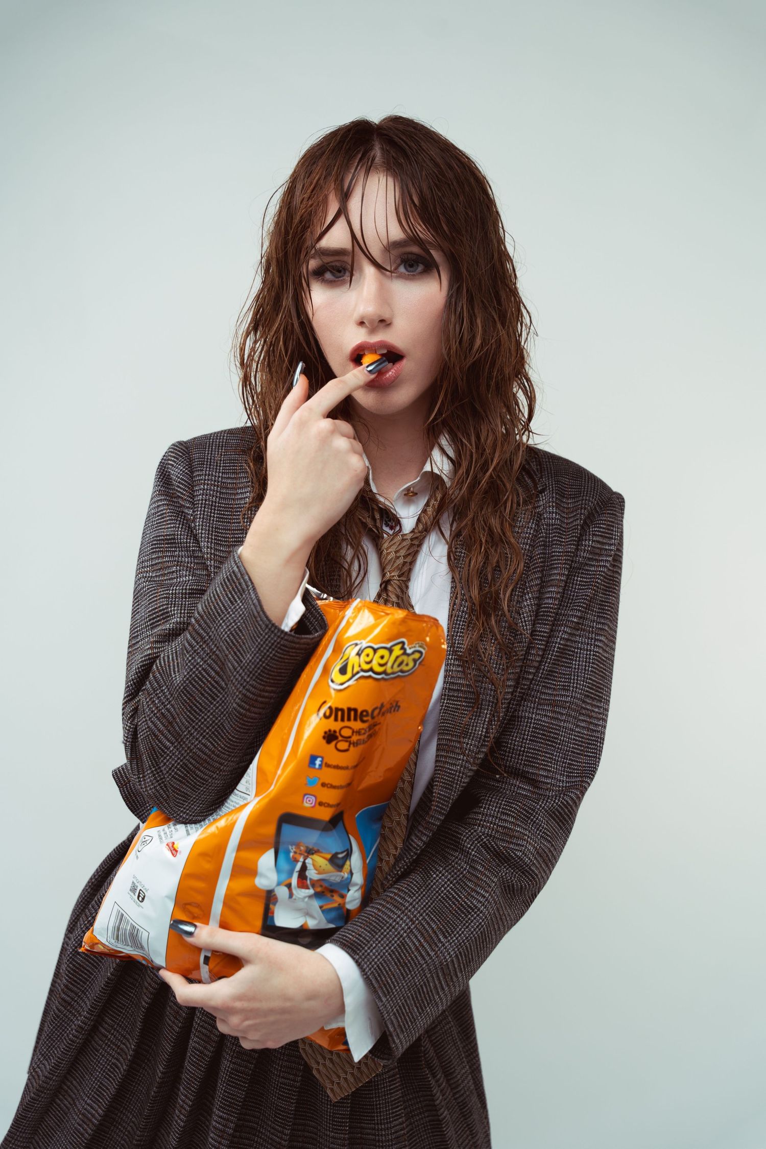 A person in a gray blazer jacket holds and eats from a bag of Cheetos while posing against a light background.