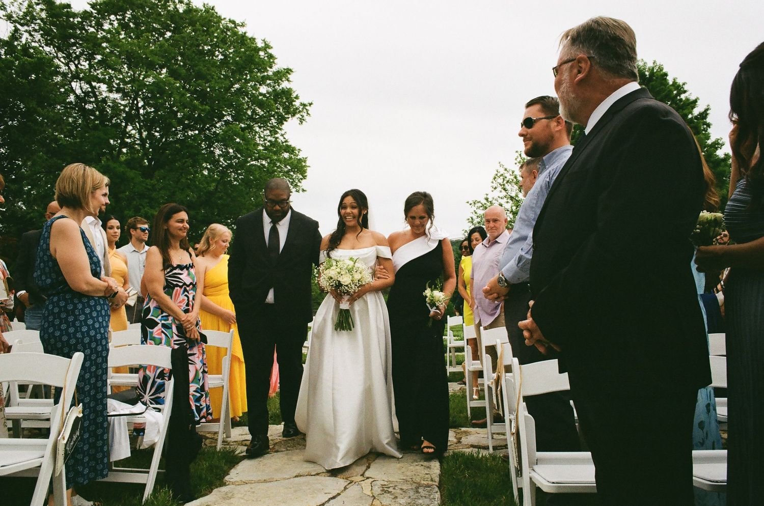 Bride walking down outdoor wedding aisle with escort, guests seated in white chairs, lush green trees in background.