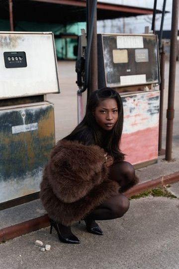 A woman in a brown fur coat and black heels crouches beside vintage gas pumps at an old station.