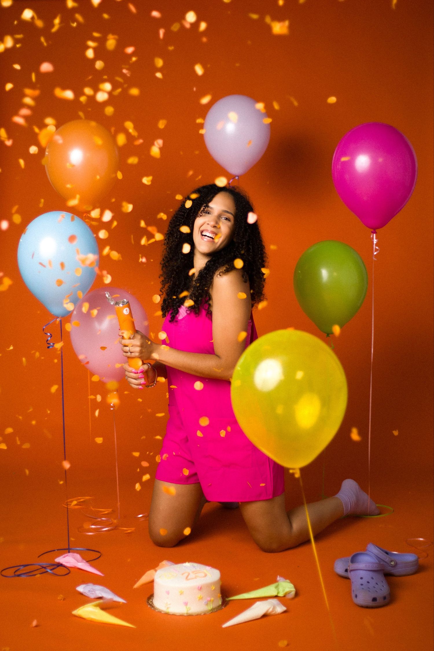 Woman in pink romper celebrating with confetti, colorful balloons, and cake on orange background.