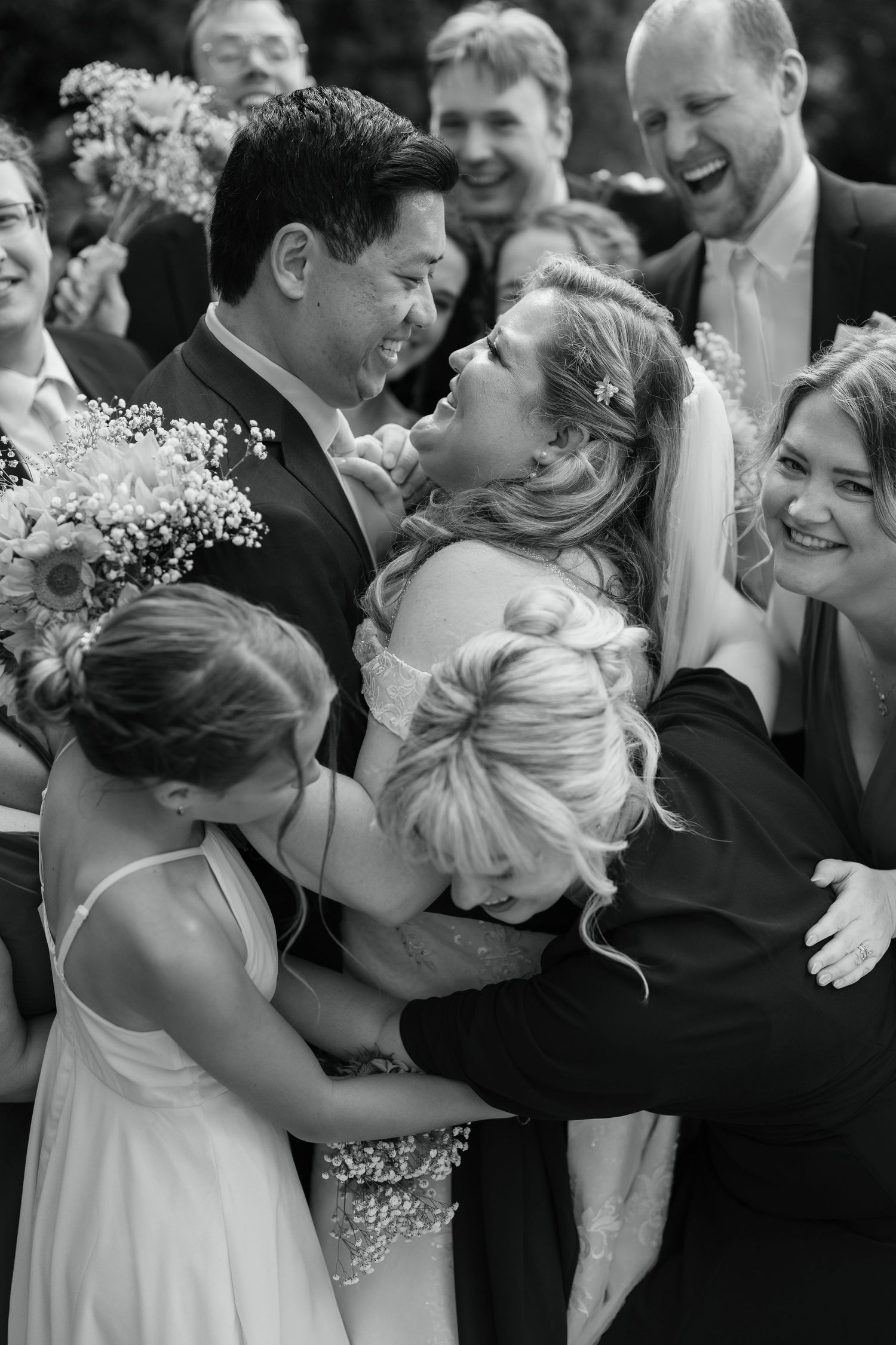 Black and white photo of a joyful bride and groom embraced by wedding guests during a celebratory group hug.