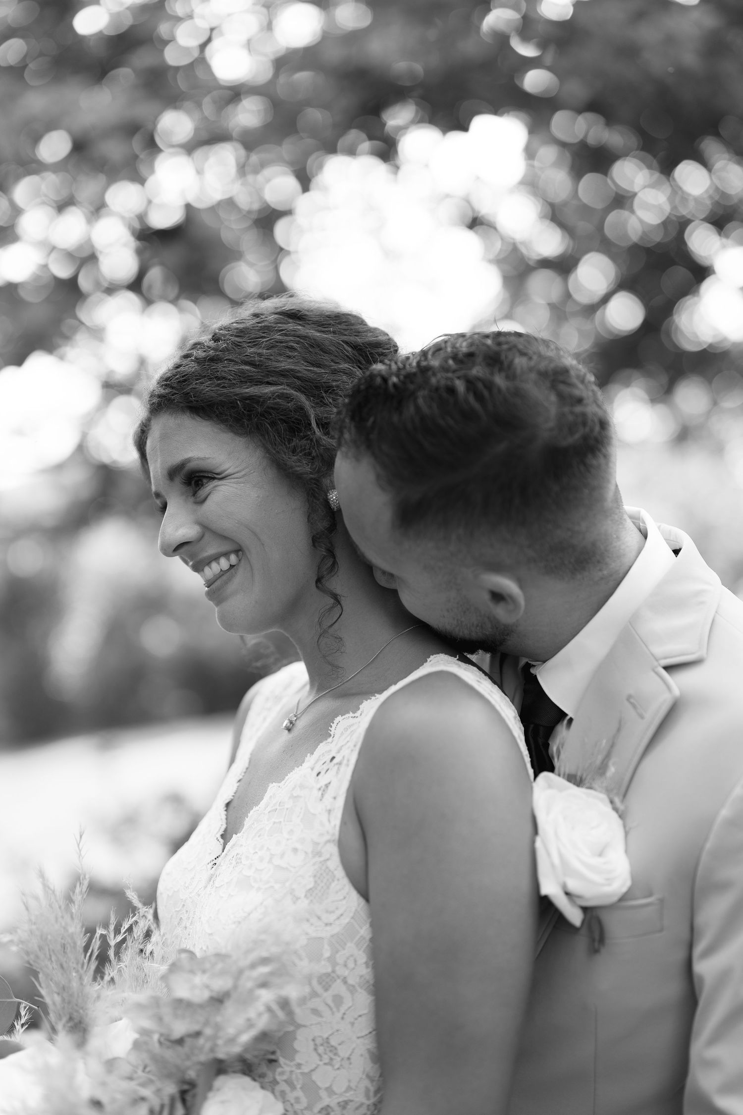 Black and white photo of a smiling bride holding flowers as groom kisses her shoulder outdoors.
