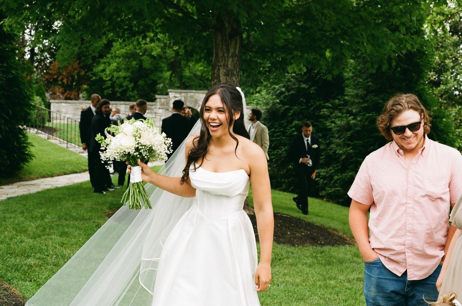 Smiling bride in white gown holding bouquet walks outdoors with guests in background at garden wedding.