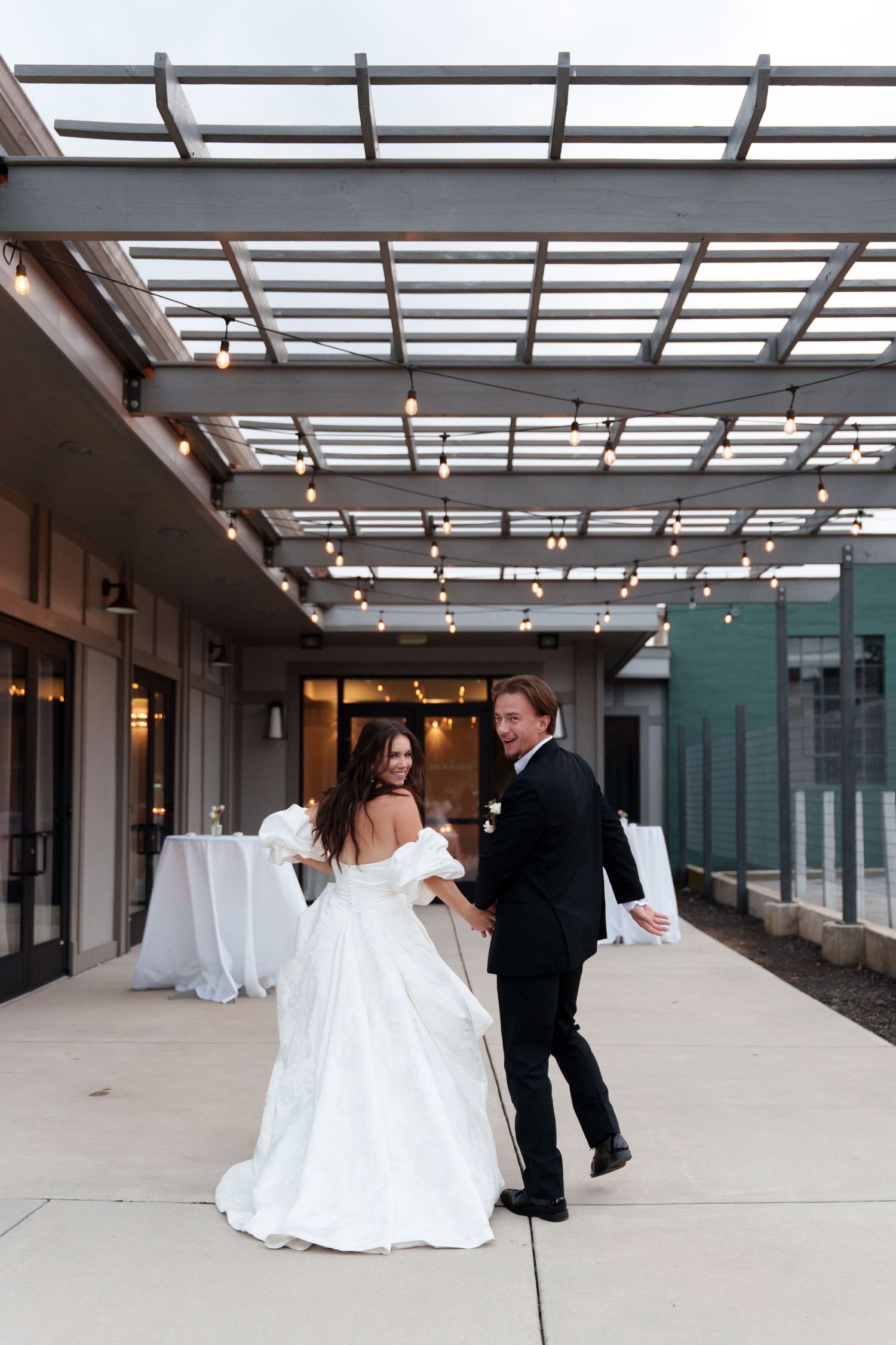A couple in wedding attire dance together beneath a modern glass-covered outdoor patio with decorative lighting.