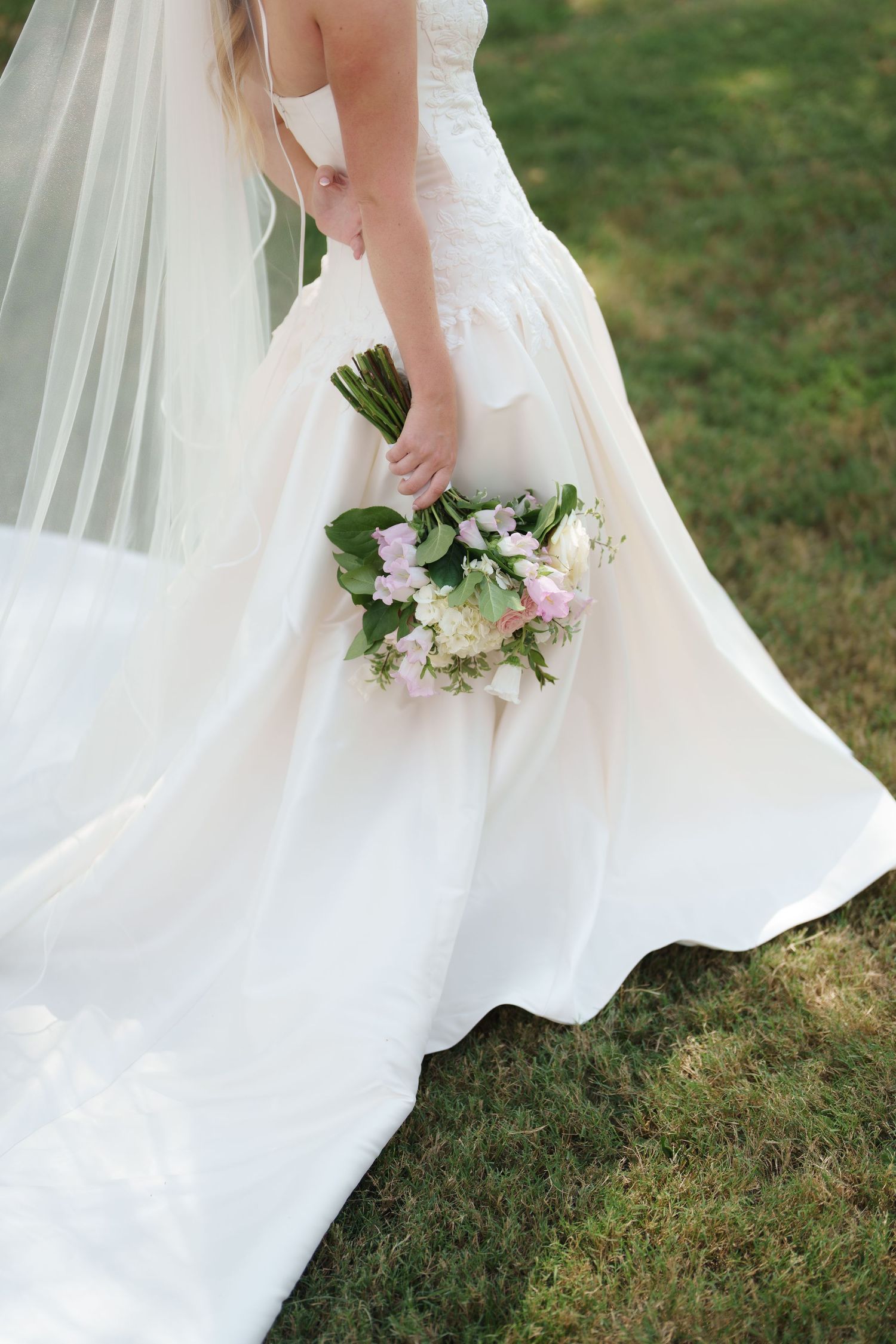 Bride in white gown holding pink and white floral bouquet with green leaves, standing on grass with flowing veil.