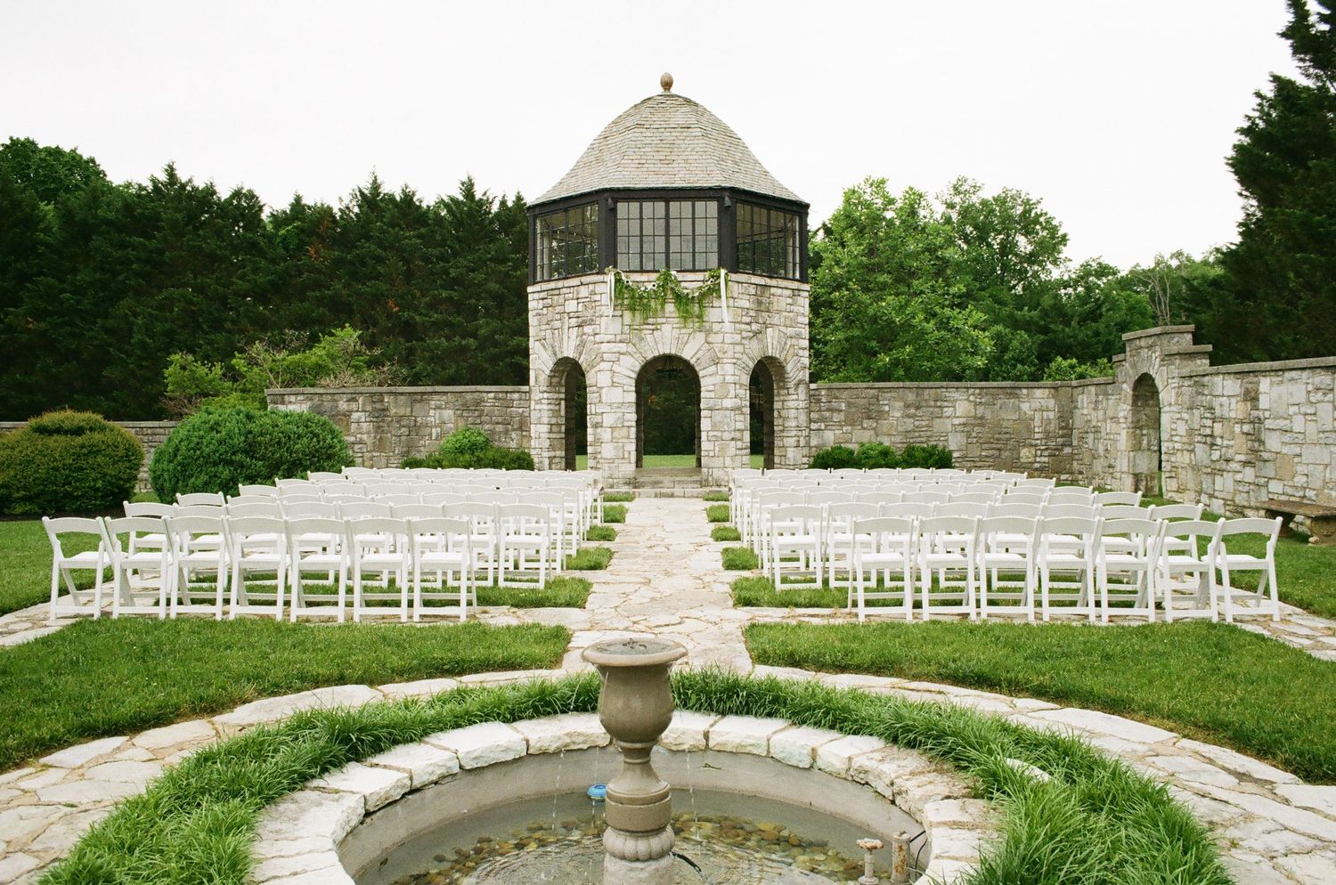 Elegant outdoor wedding ceremony setup with white chairs, stone walls, fountain, and a historic gazebo surrounded by lush greenery.