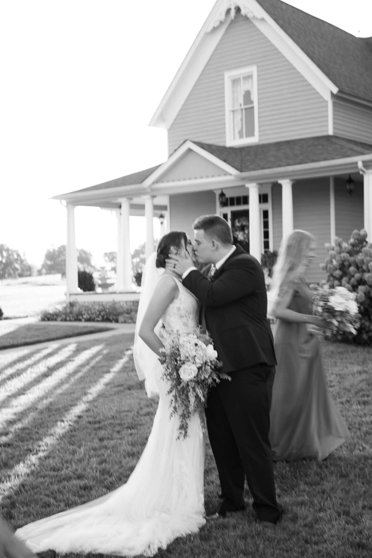 Black and white photo of a bride and groom sharing a kiss outside a charming farmhouse on their wedding day.