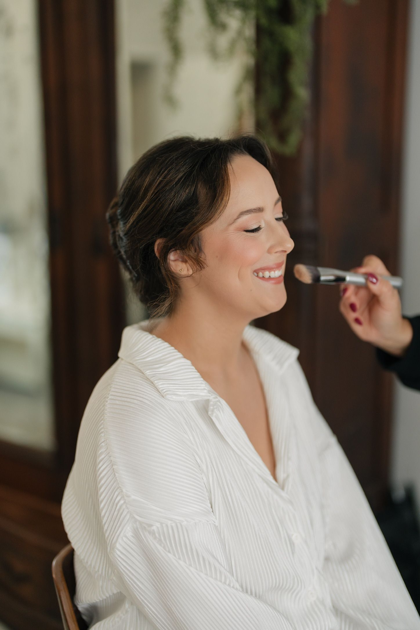 Smiling bride getting makeup applied with a brush while wearing a white striped robe during wedding preparations.