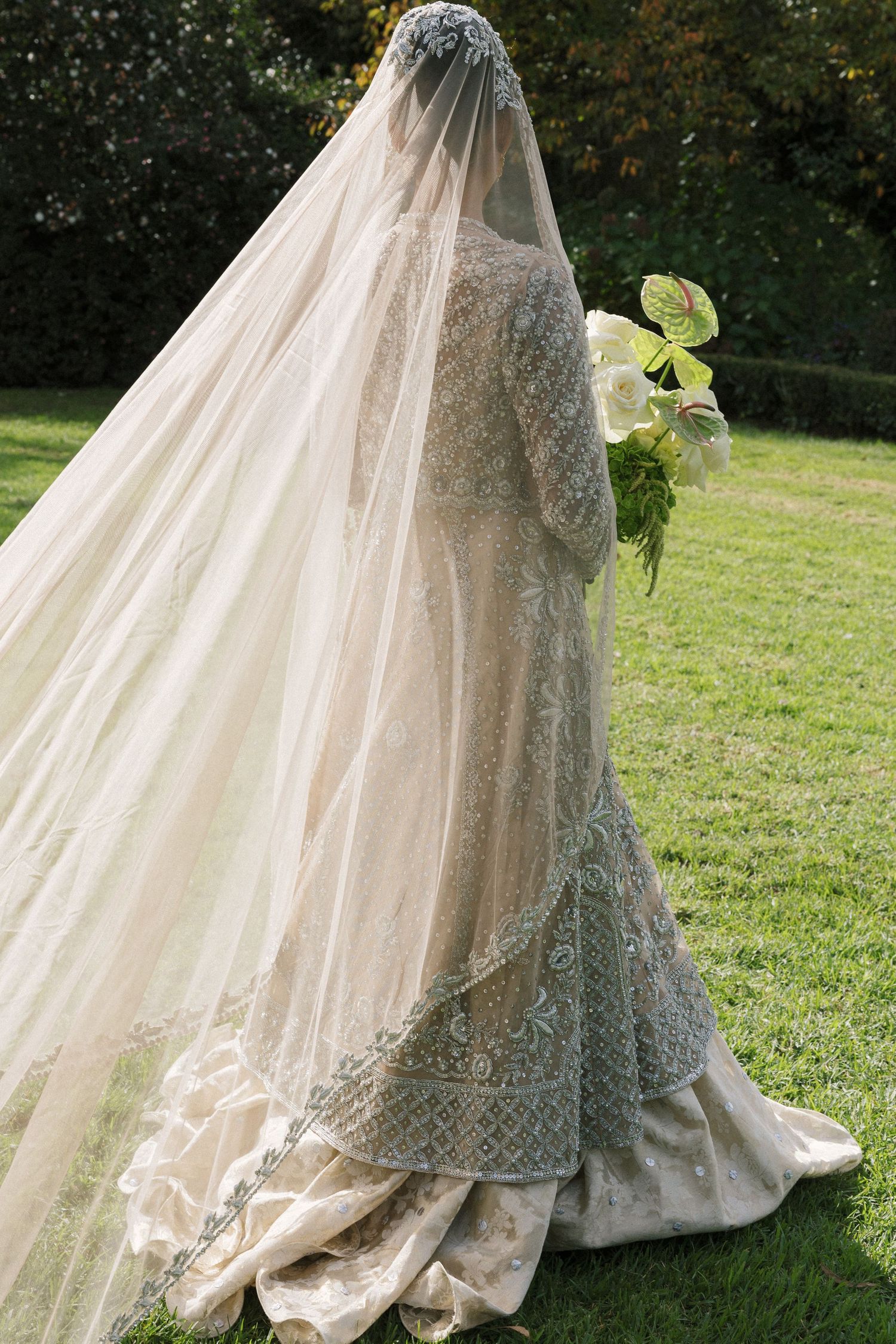 Bride in embroidered sage green lehenga with flowing ivory veil holds white bouquet outdoors on lush green lawn.