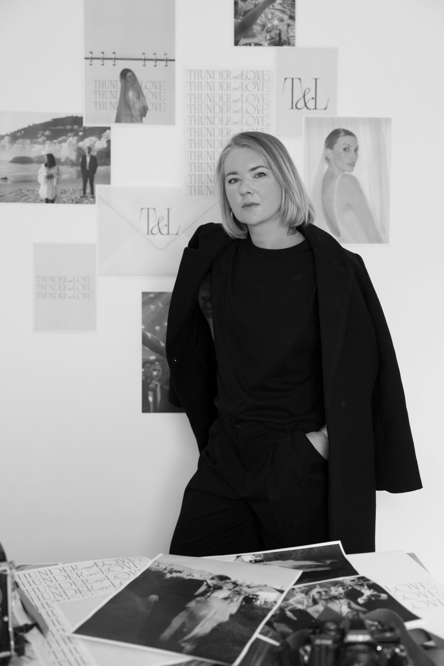 A young woman with blonde hair wearing a black outfit stands in front of a wall with various photographs displayed.