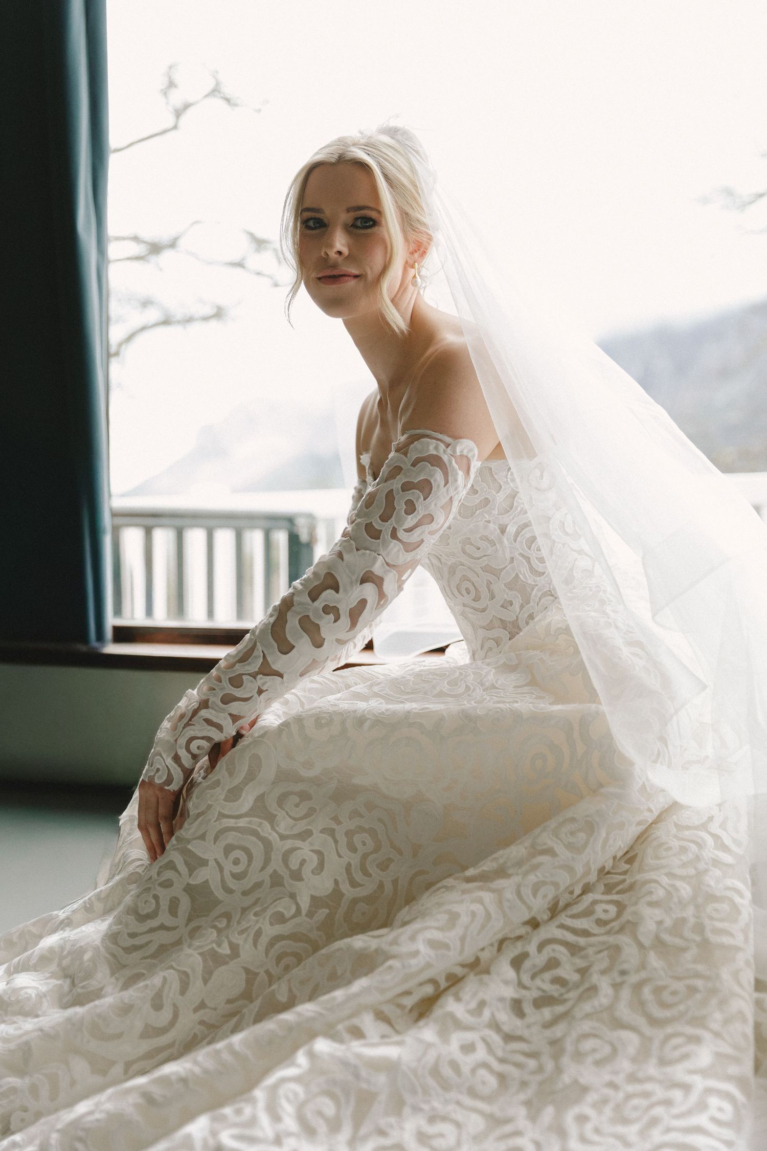 A bride in an elegant lace wedding gown poses gracefully by a window in a series of romantic bridal portraits.