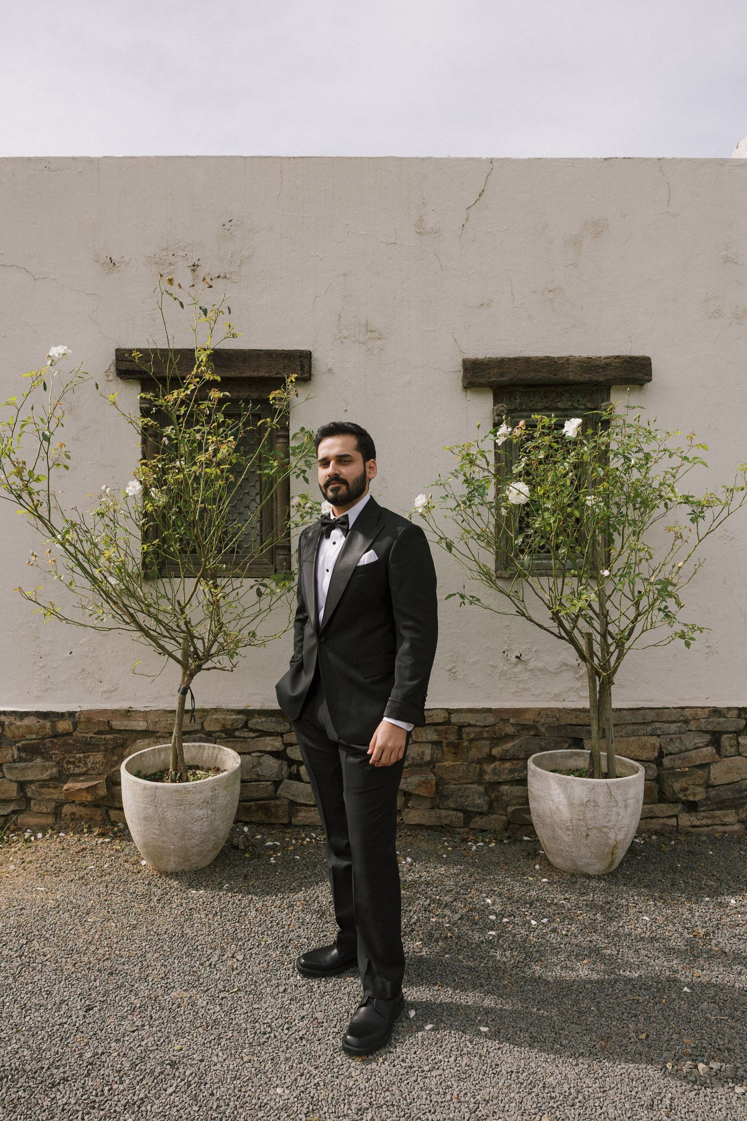 A dapper groom in a black tuxedo with bow tie stands between two potted trees outside a white stucco building.