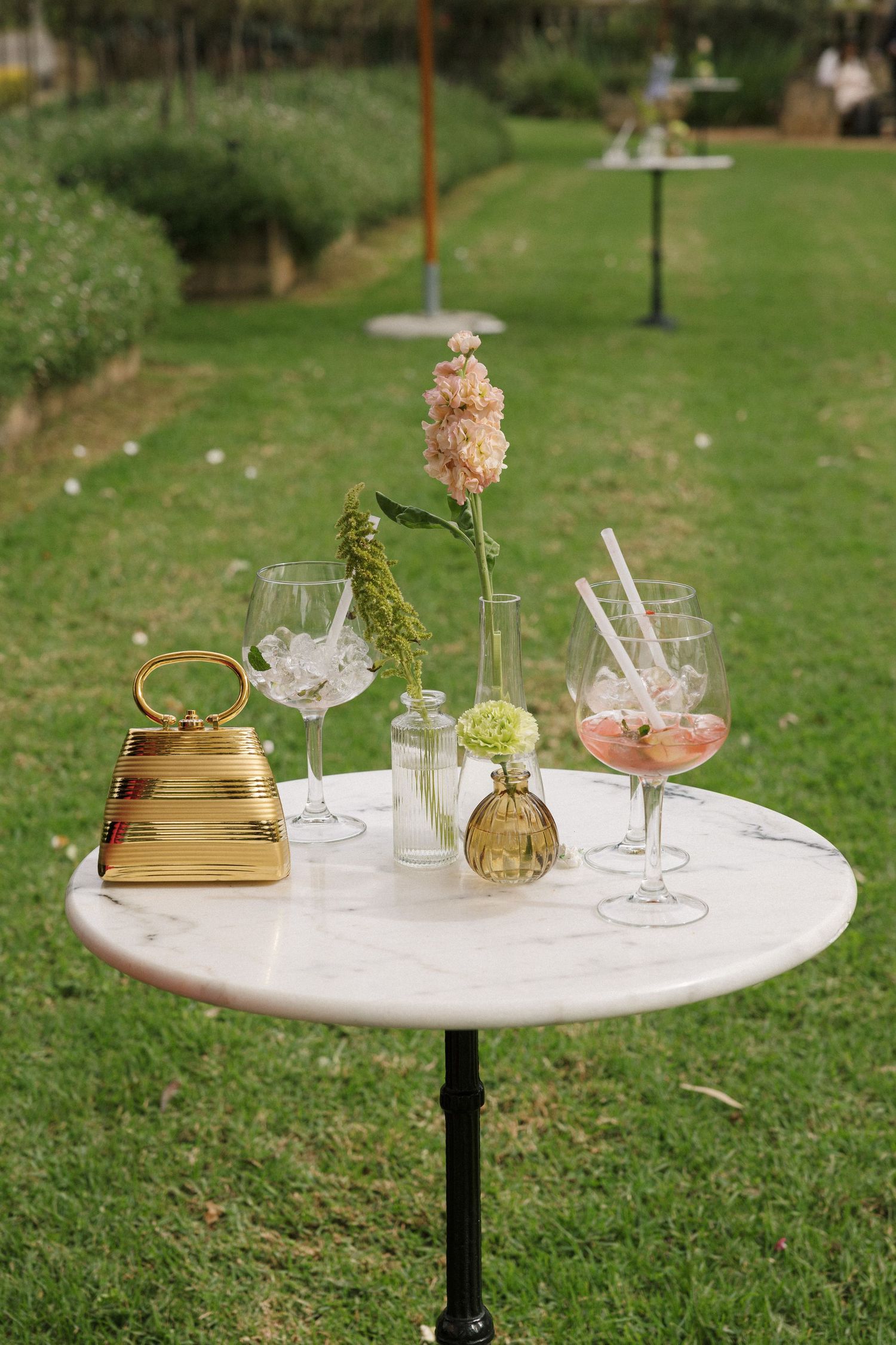 Outdoor garden cocktail table with wine glasses, pink drink, gold handbag, and fresh flowers on marble top.