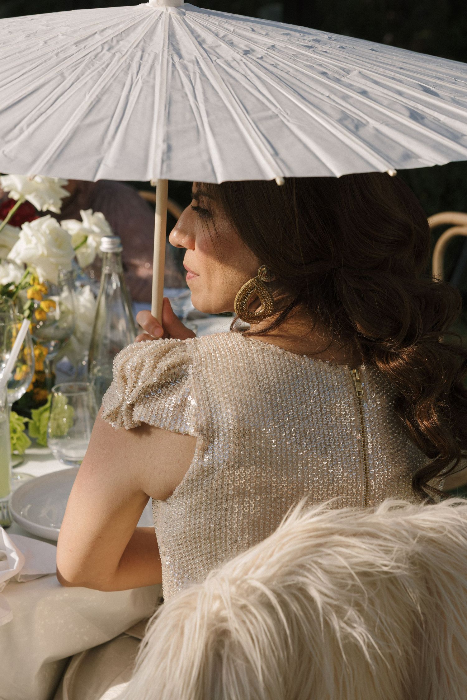 Woman in sequined dress holding white parasol umbrella seated at elegant outdoor table with floral arrangements.