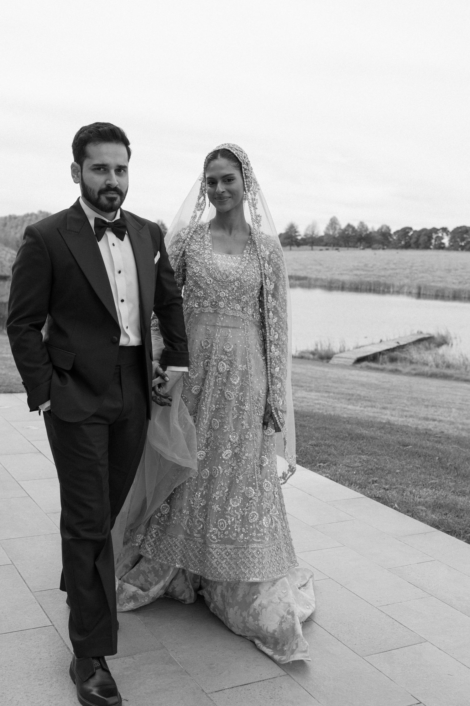 Black and white photo of a bride in embroidered lehenga with veil and groom in tuxedo walking hand in hand outdoors.