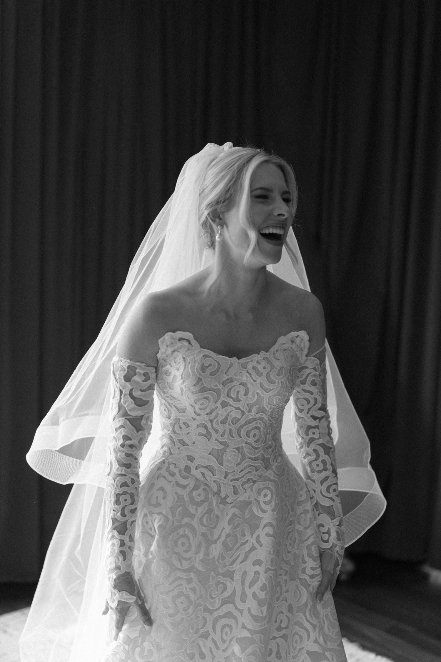 Black and white photo of a bride in an ornate lace wedding gown with long sleeves and flowing veil.