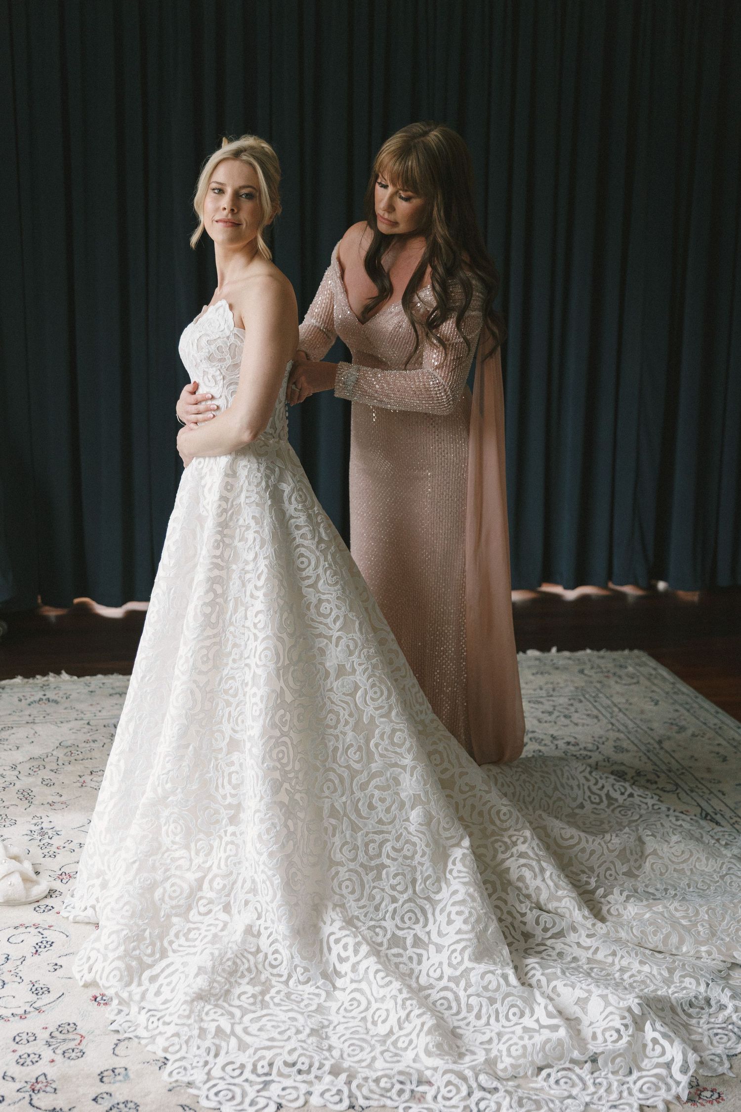 Two people in formal attire stand together, one wearing an intricate white lace wedding gown with a long train.