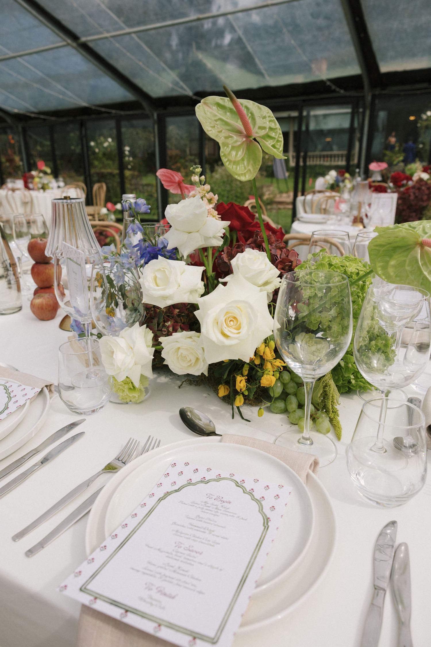 Wedding reception table with white roses, anthurium centerpiece, menu card, and crystal glassware in greenhouse.