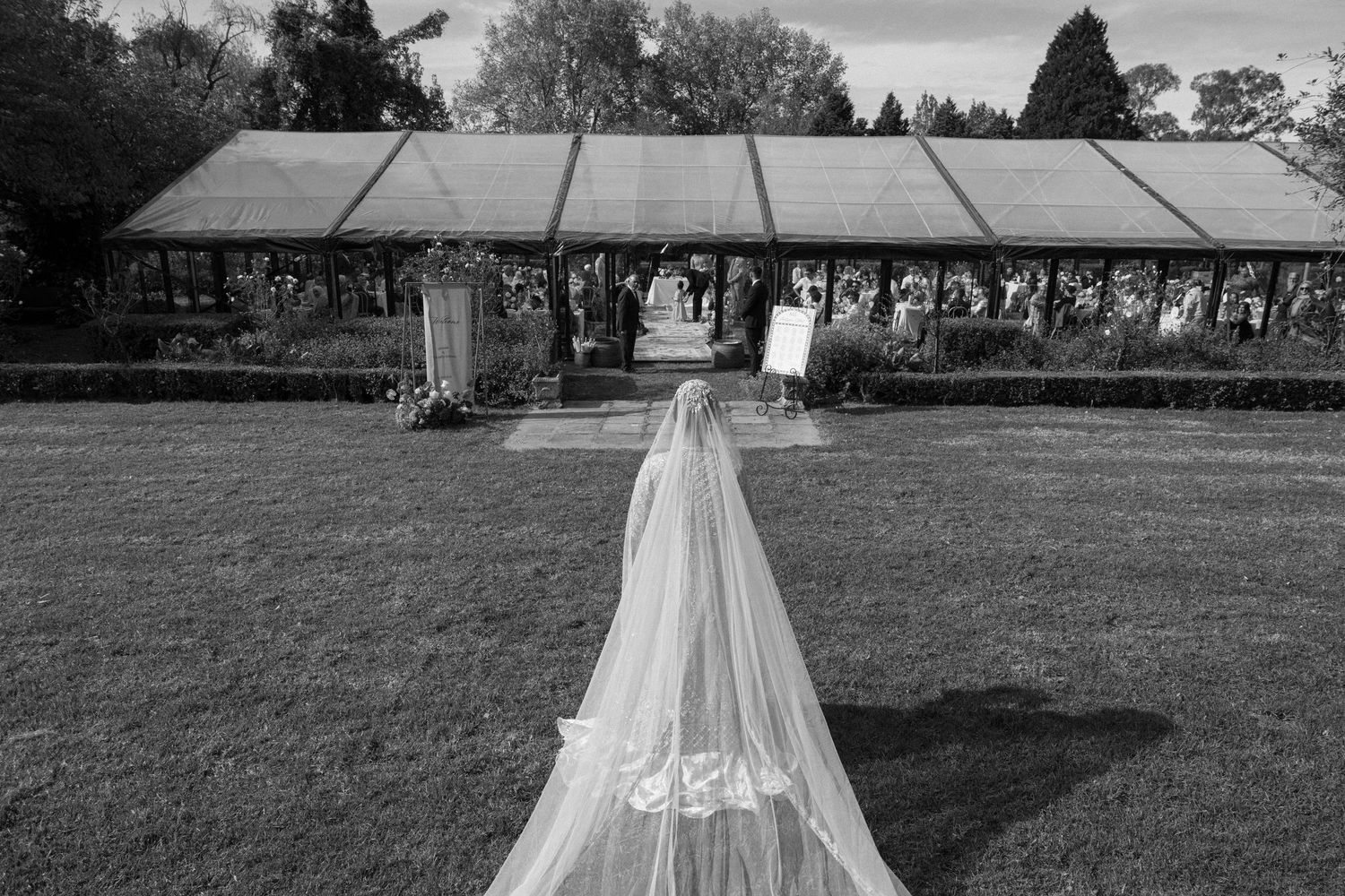 Black and white aerial view of bride's long wedding veil trailing across lawn toward outdoor ceremony tent.