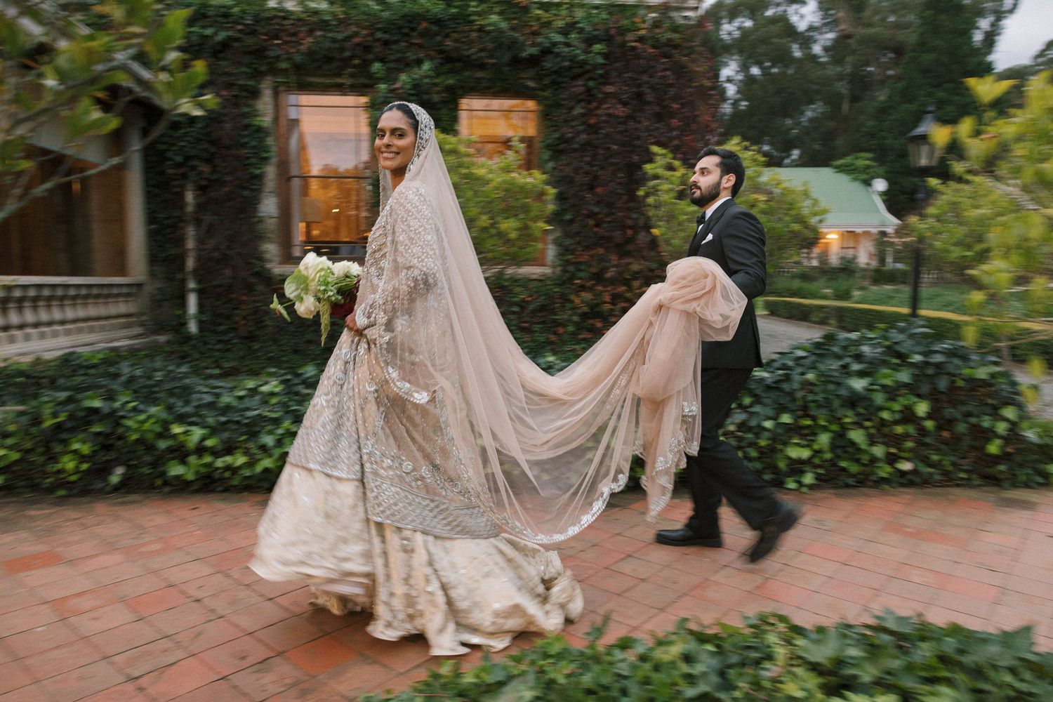 Bride in ornate South Asian wedding gown with long veil walks with groom through ivy-covered garden venue.
