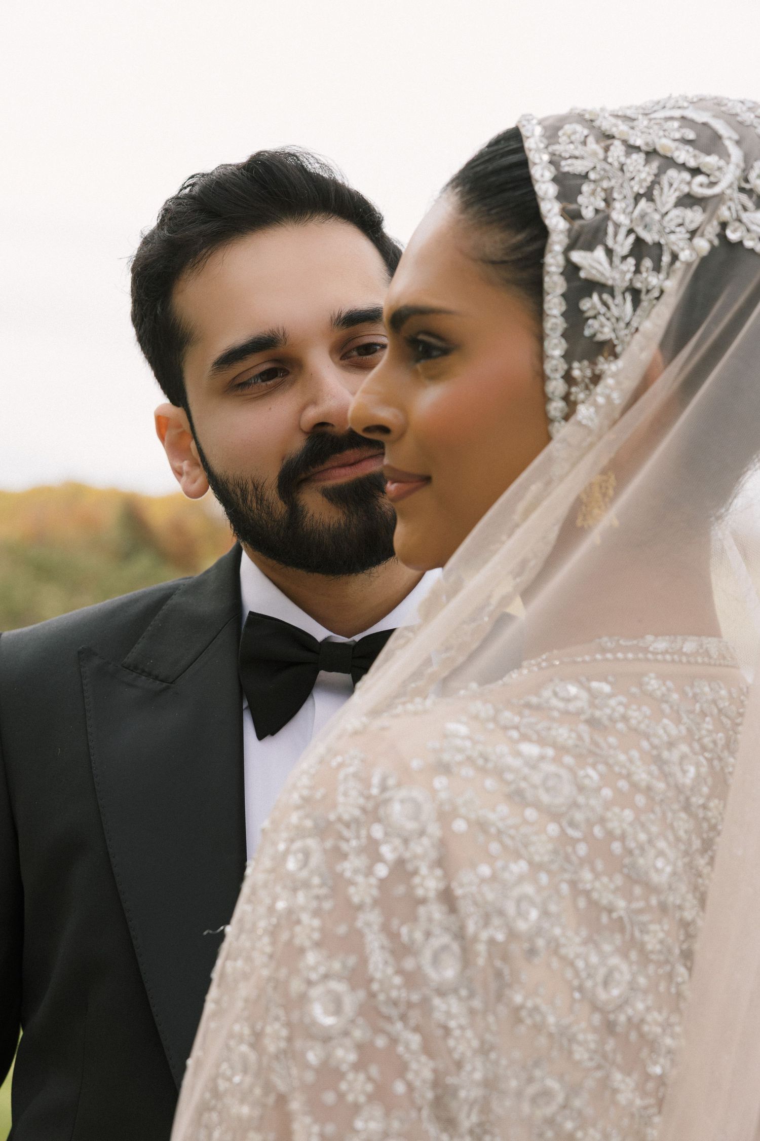 A bride in a beaded white gown and jeweled veil stands close with a groom in a black tuxedo outdoors.