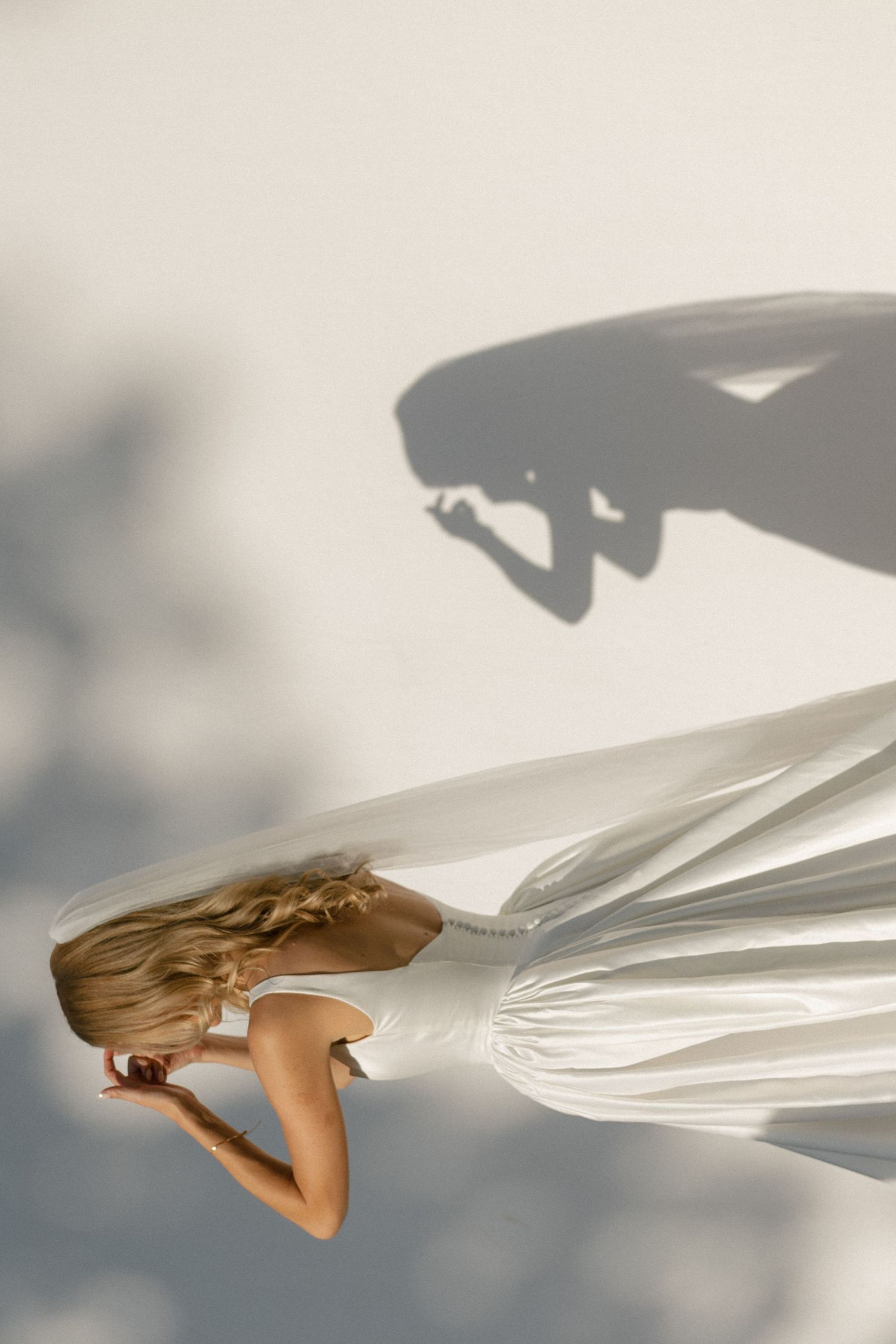 Artistic photo of a person in a flowing white dress jumping against a minimalist background with dramatic shadows.
