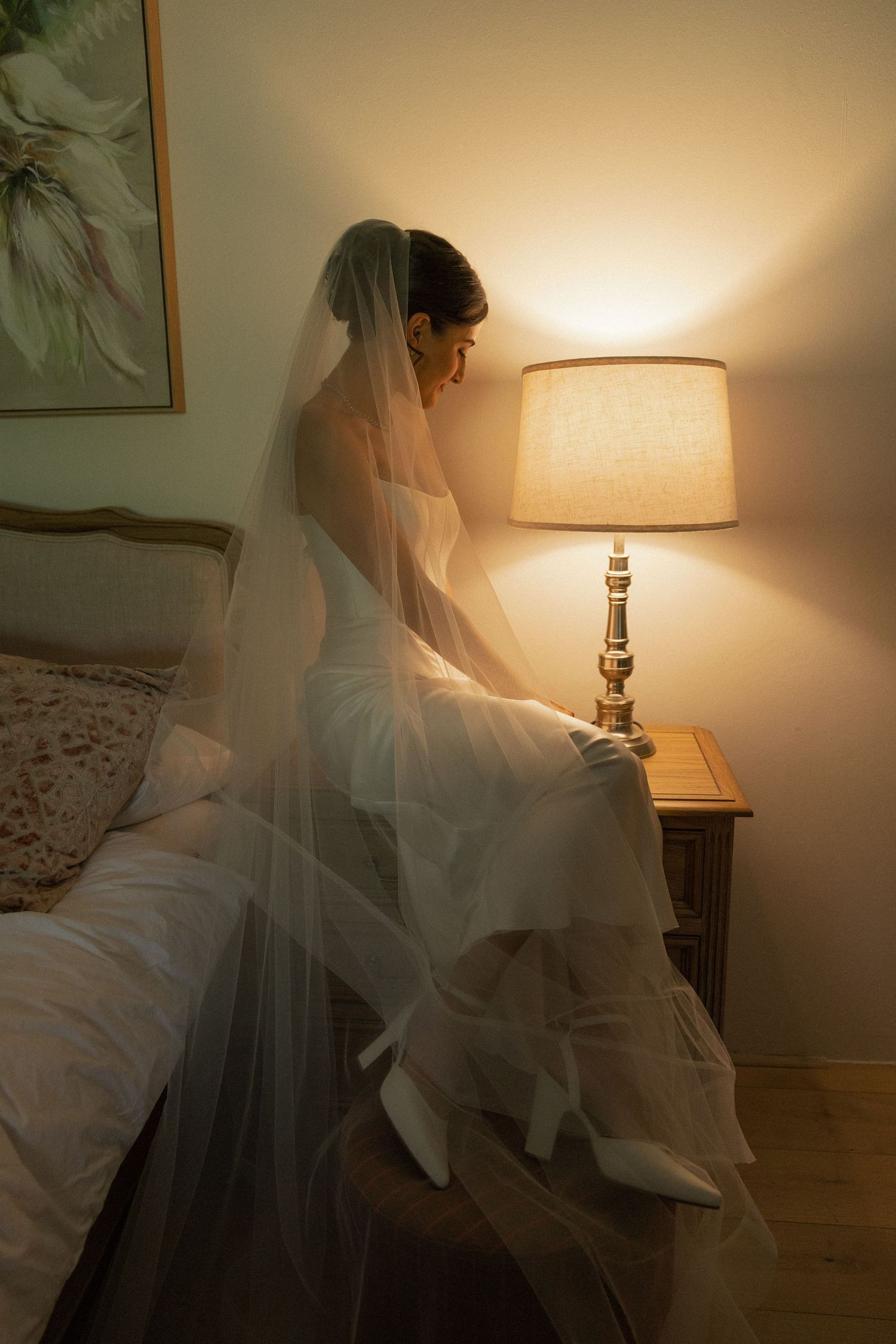 Bride in white wedding gown and veil sits on bed beside glowing lamp in softly lit bedroom.