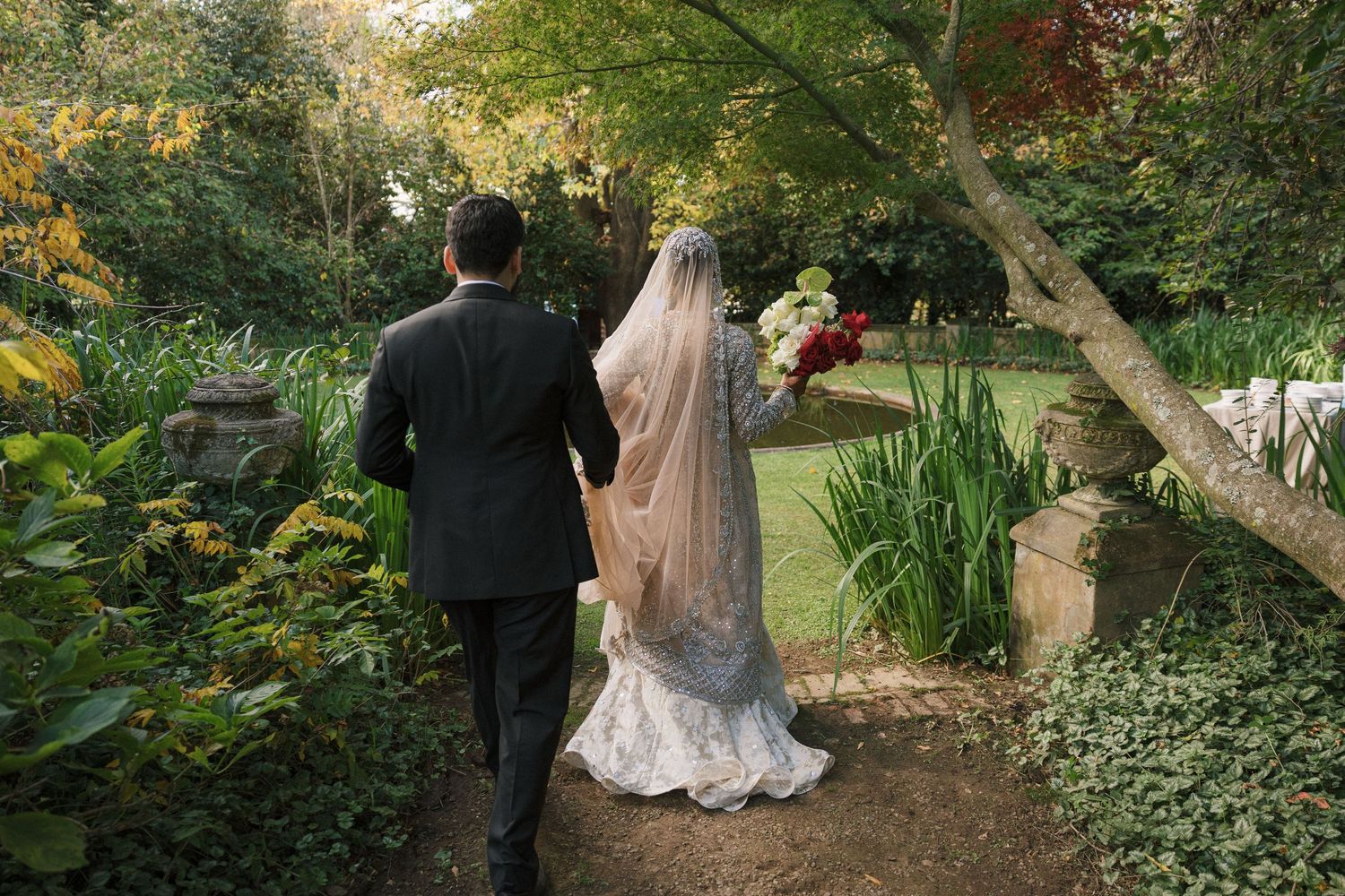 Bride and groom walking hand in hand through a lush garden path, bride wearing white gown with veil holding red bouquet.
