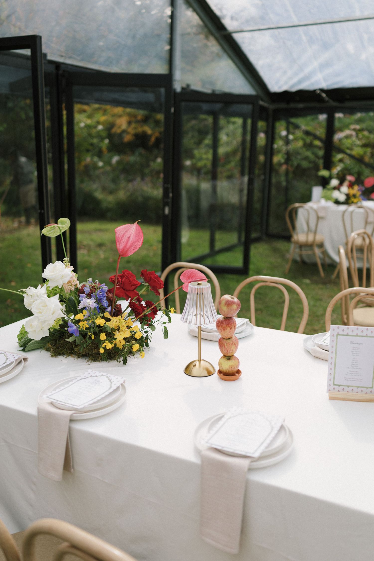 Elegant wedding reception table with white linen, colorful floral centerpiece, and rattan chairs inside a glass greenhouse.