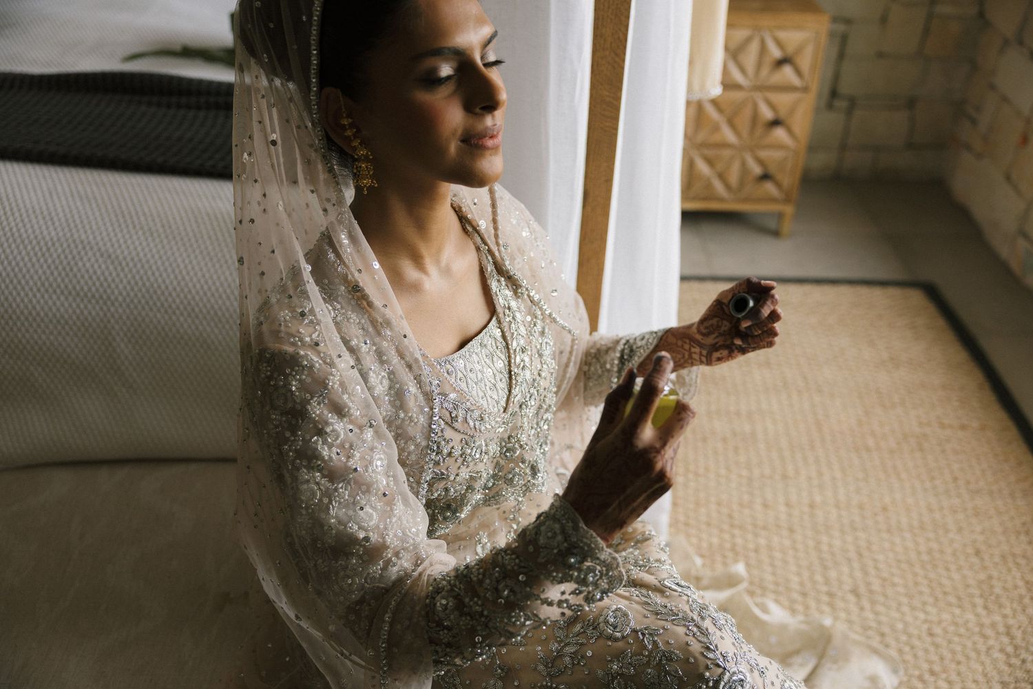 Bride in ornate white lace bridal gown and veil sits gracefully holding perfume bottle near window.