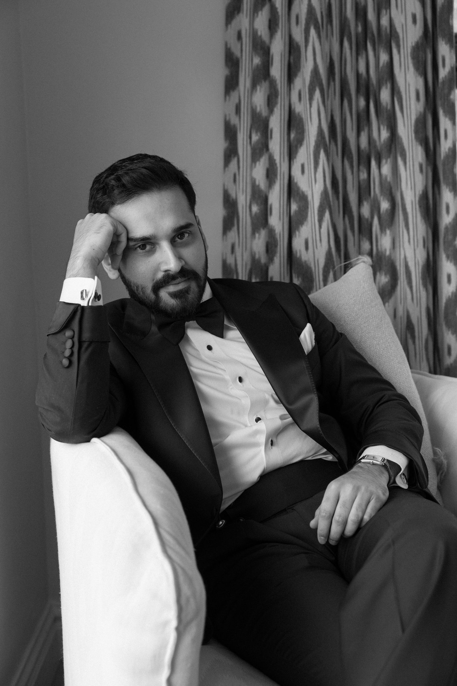Black and white portrait of a bearded man in a tuxedo sitting relaxed in a chair, resting his head on his hand.