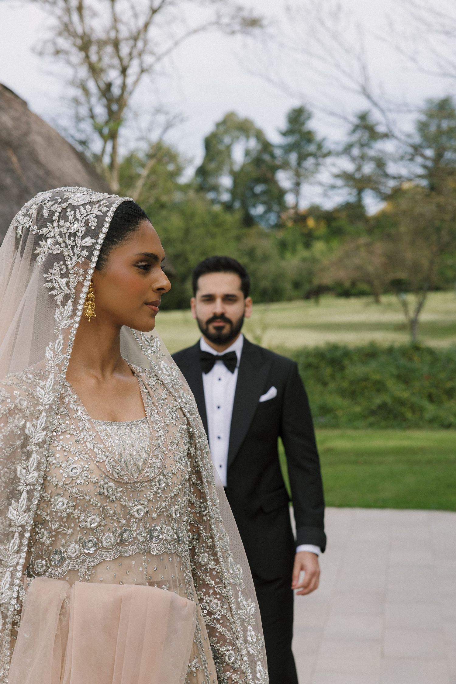 South Asian bride in embroidered lehenga and lace dupatta with groom in black tuxedo at outdoor wedding.