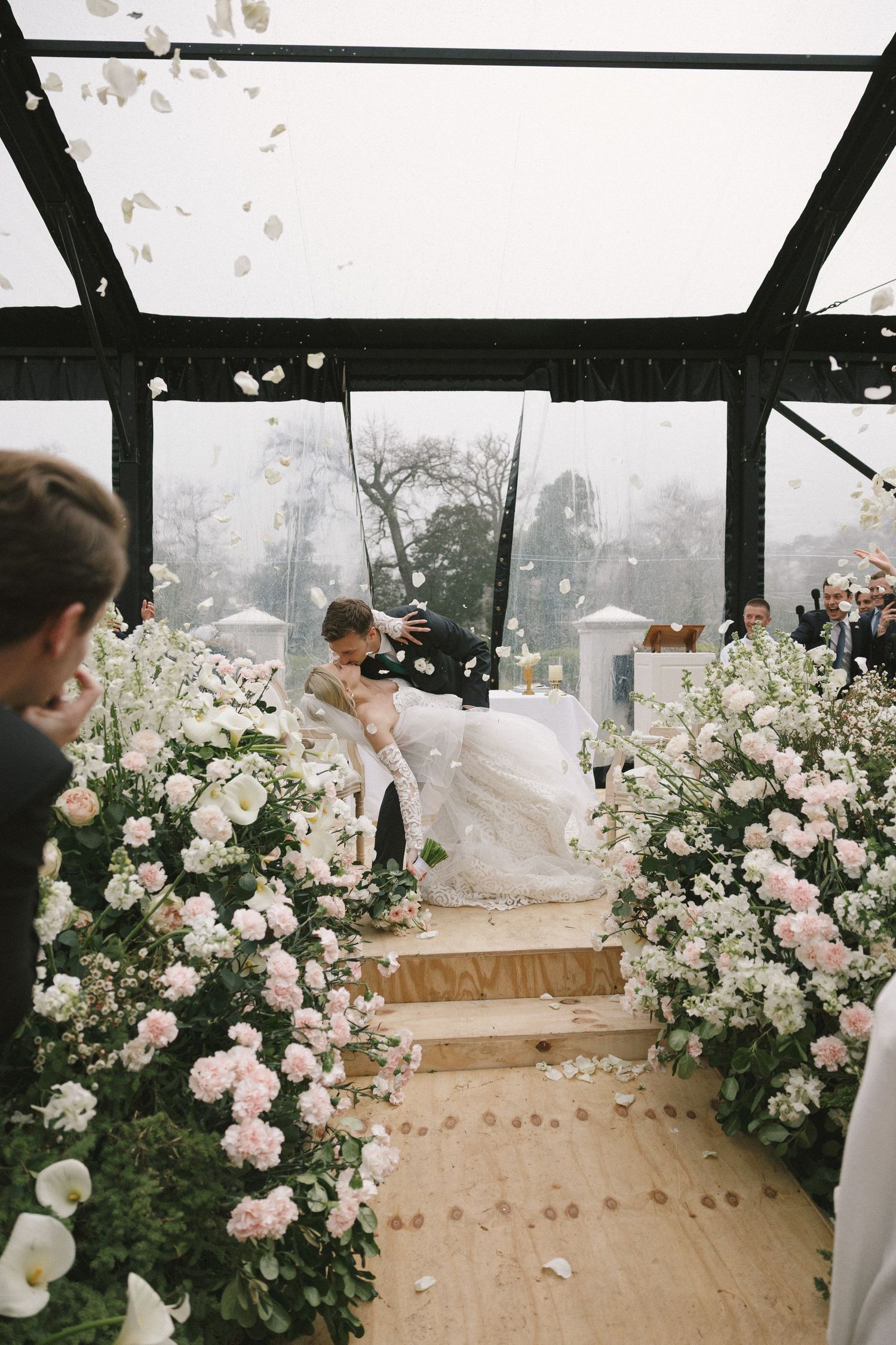 A panoramic view of a wedding ceremony setup with blush pink and white floral arrangements under a black pergola.