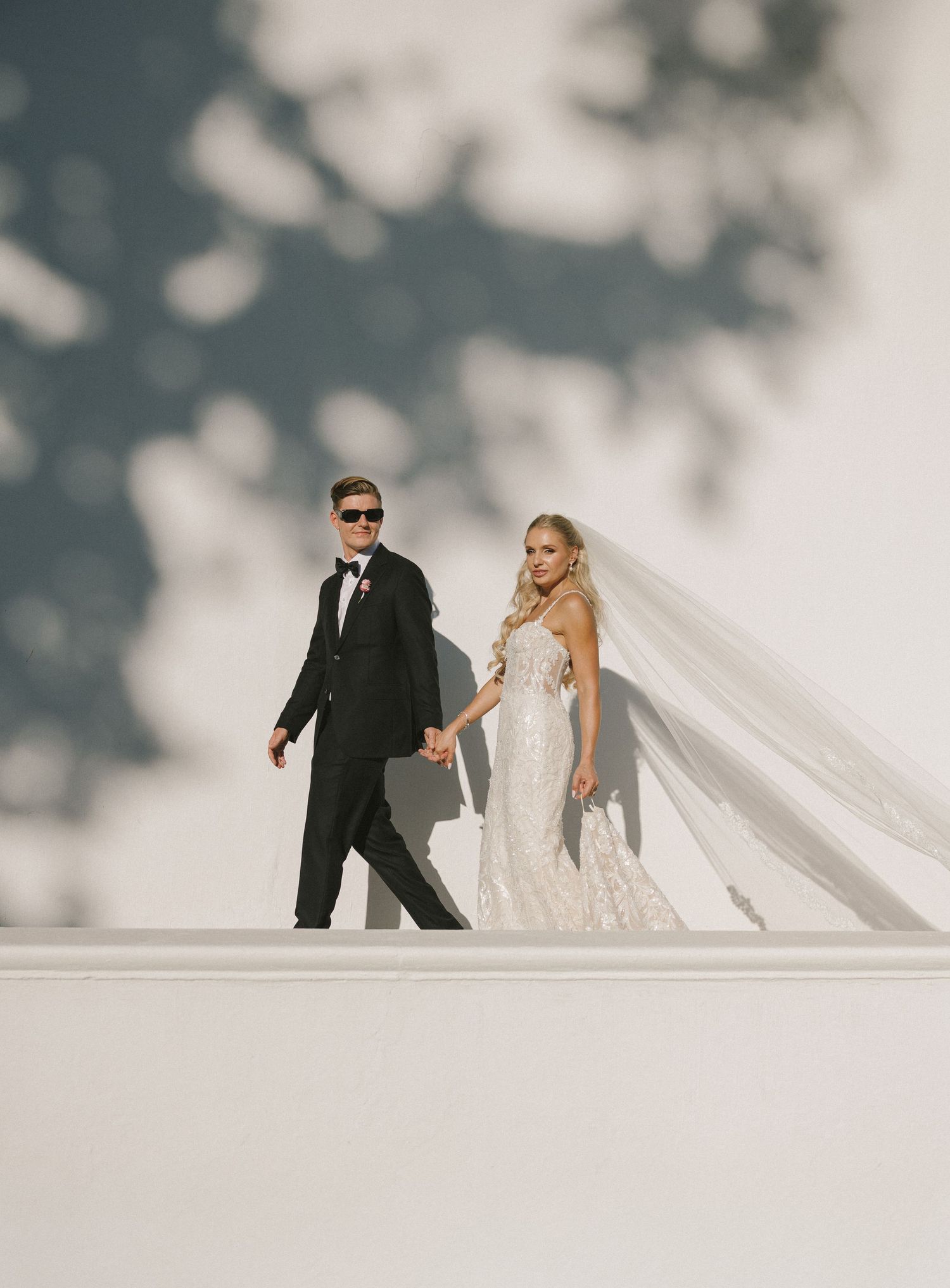 A bride and groom stand together in a formal wedding attire against a blurred natural backdrop.
